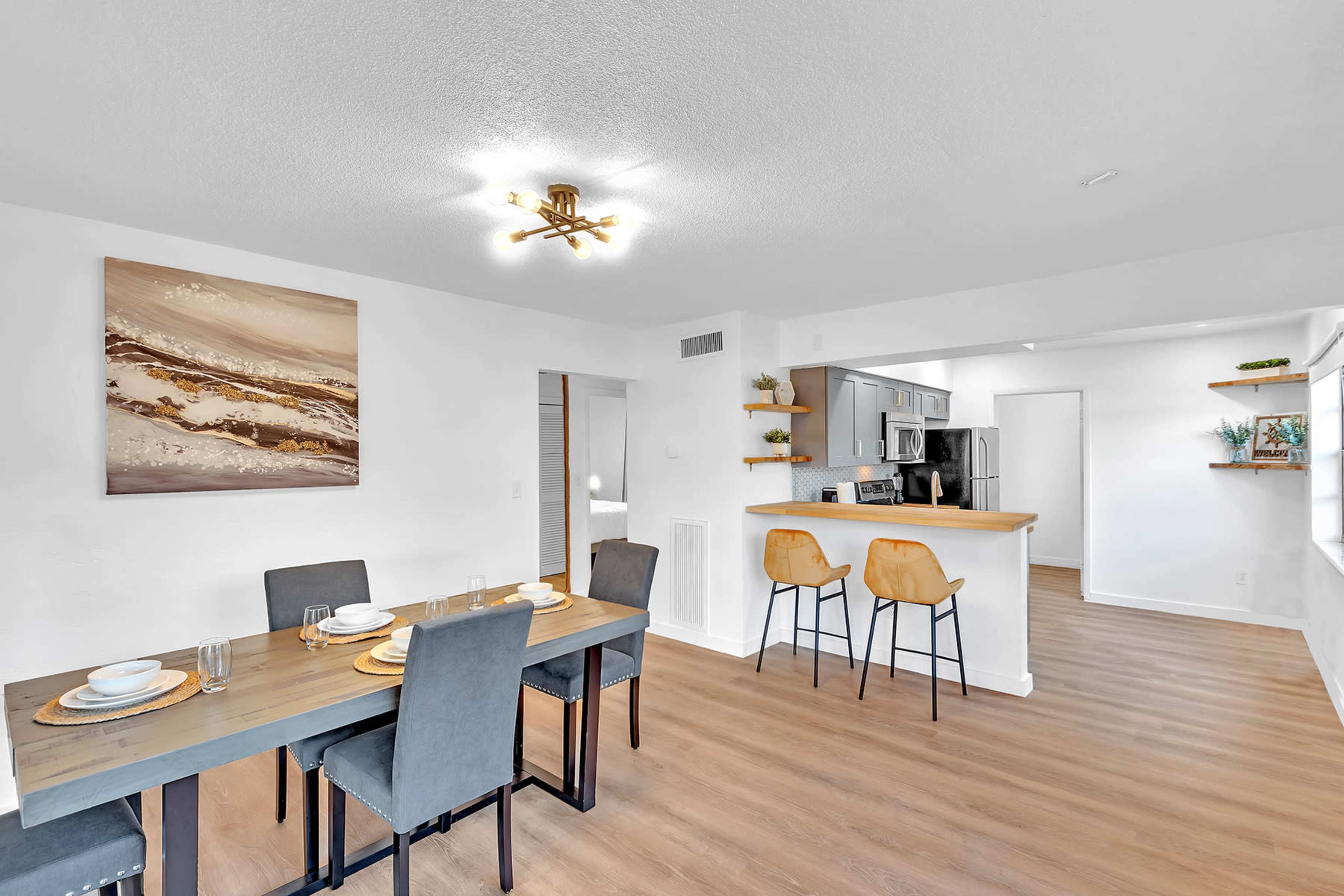 A modern dining area with a wooden table set for four, adjacent to an open kitchen with bar seating.