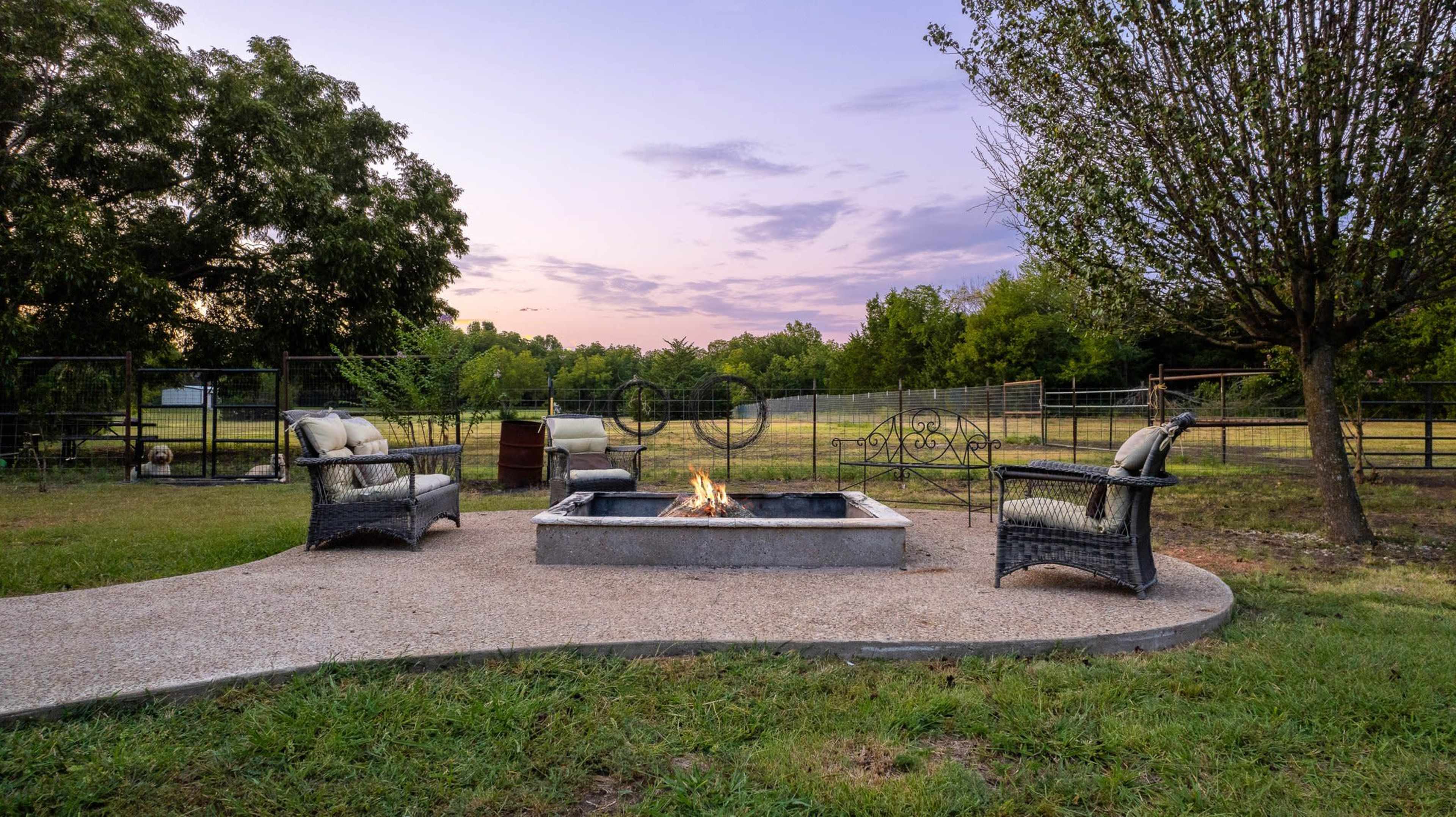 A fire pit surrounded by two chairs sits on a stone patio in an open yard with trees and a fenced area in the background.