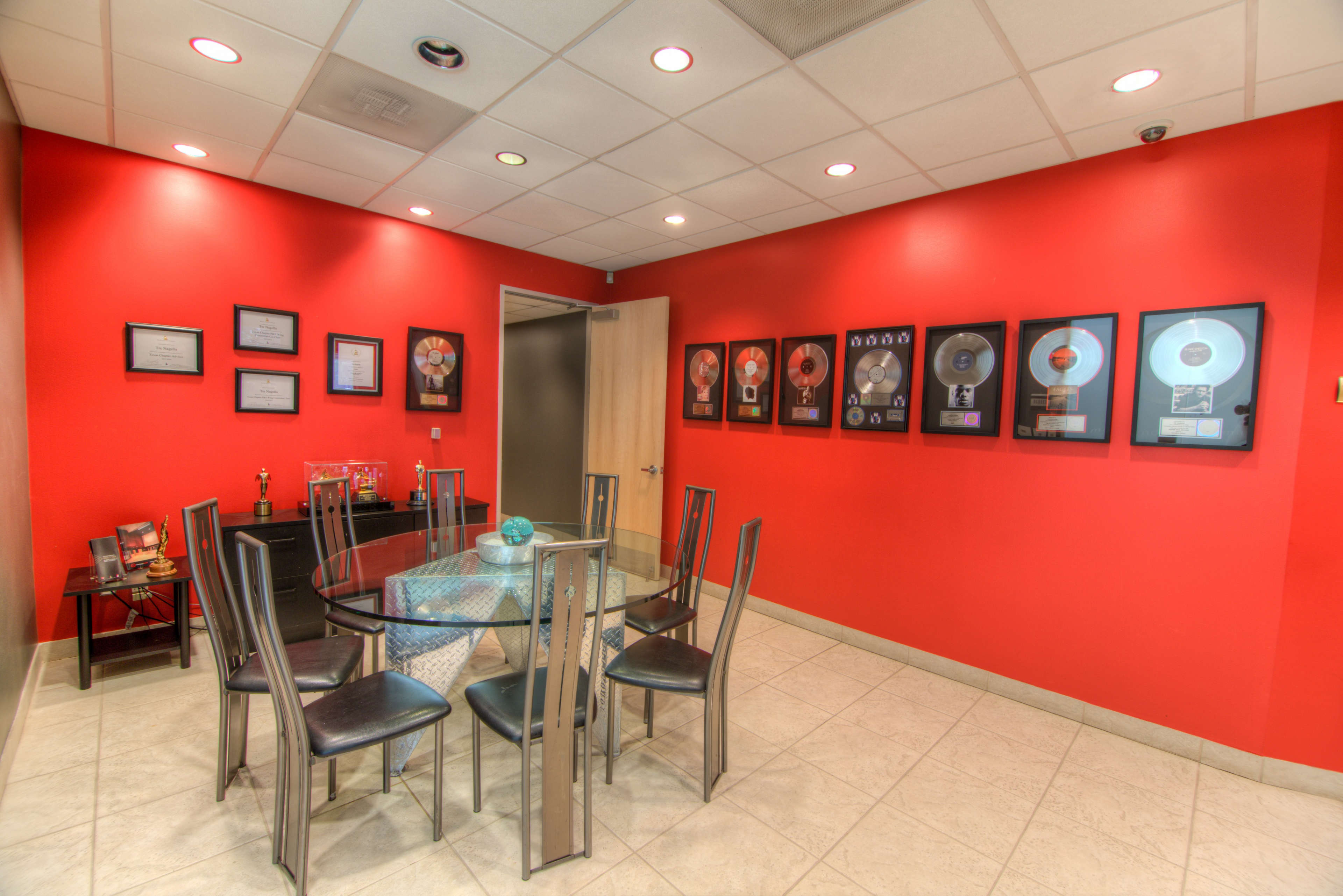 A conference room featuring a glass table surrounded by black chairs, with red walls displaying framed music awards and certificates.