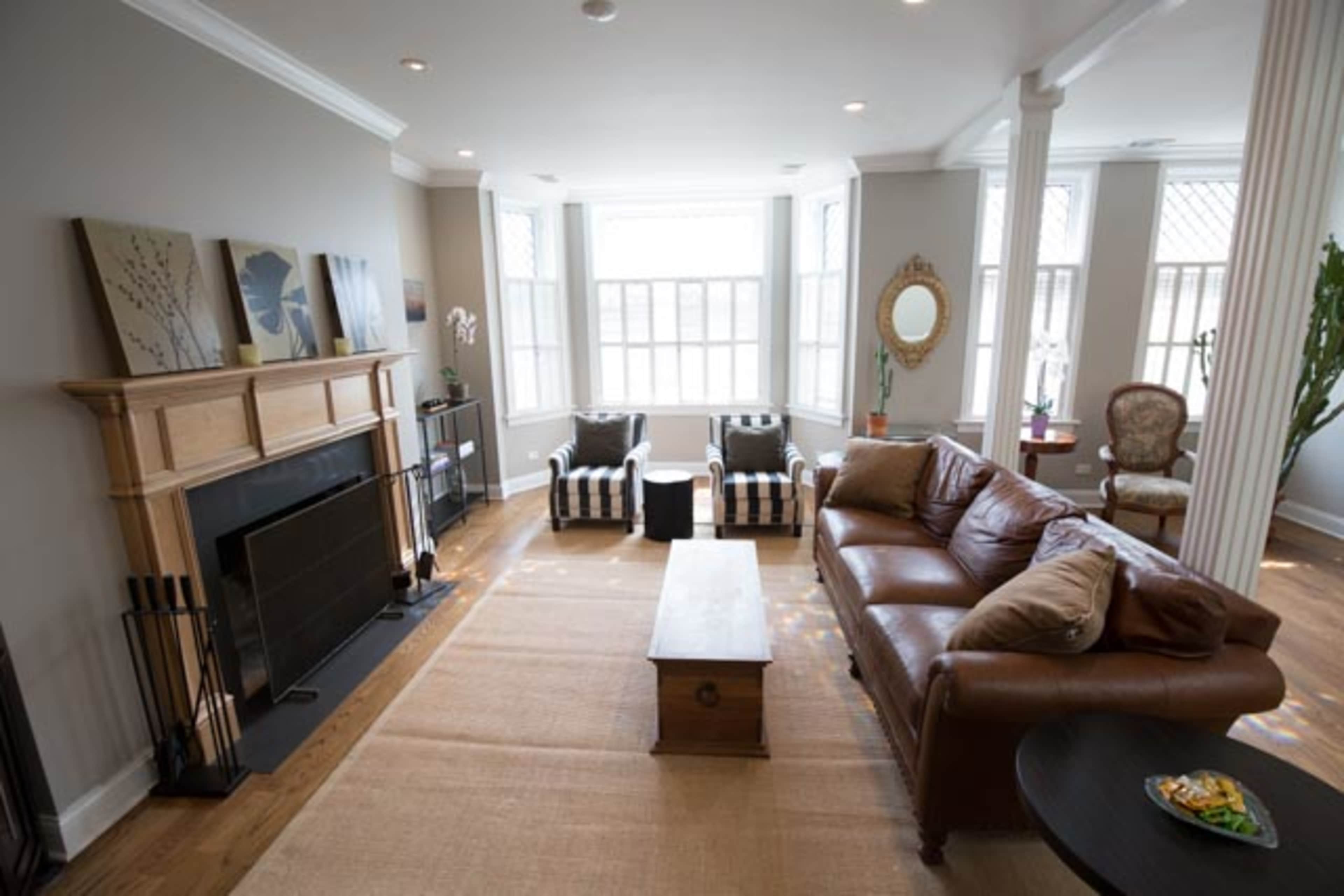 The image shows a living room featuring a leather sofa, striped armchairs, a fireplace, and large windows allowing natural light.