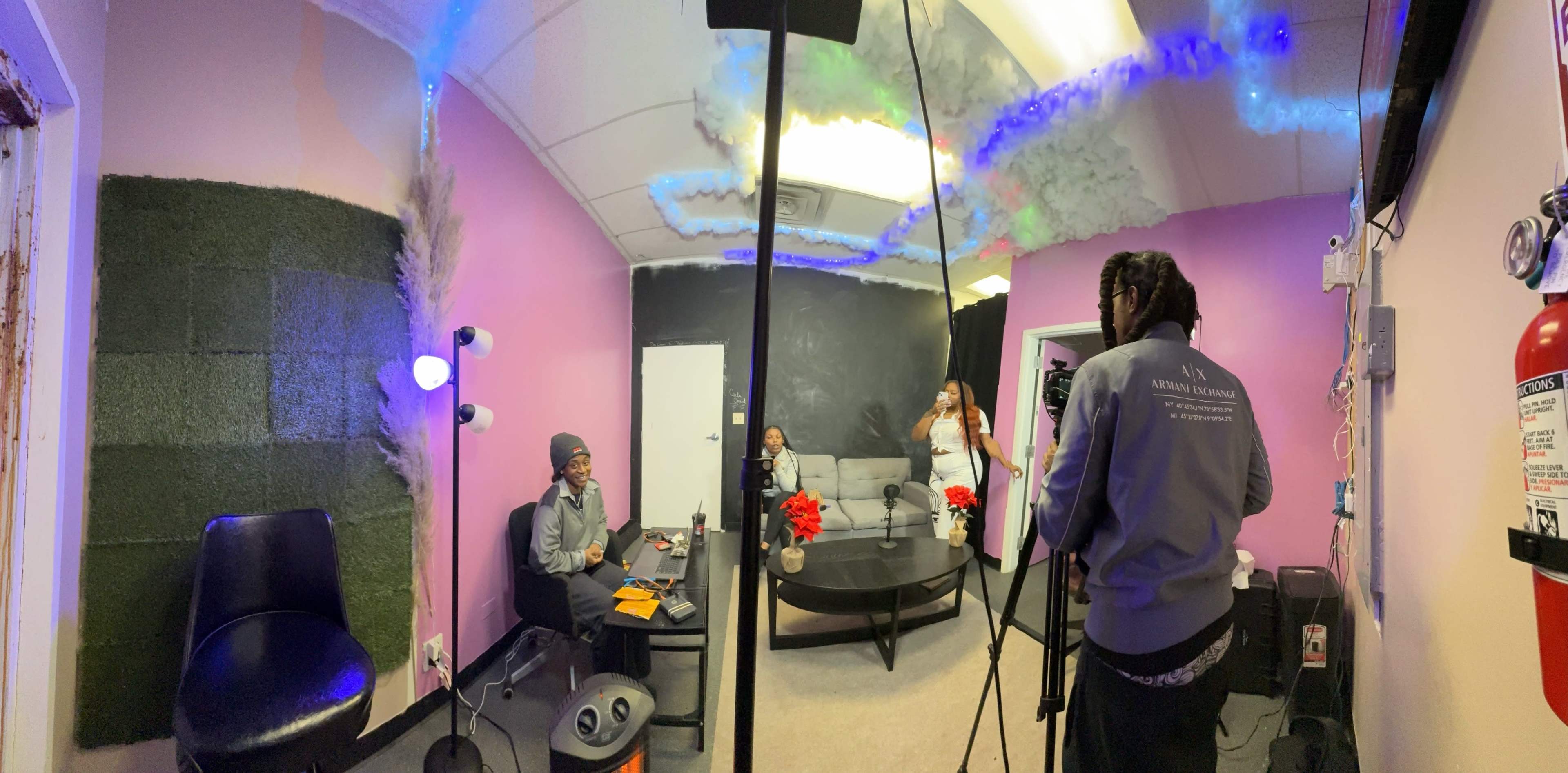 A recording session is taking place in a small studio with pink walls, a black ceiling, and a sofa, where two people are seated at a table while another person stands nearby holding a camera.