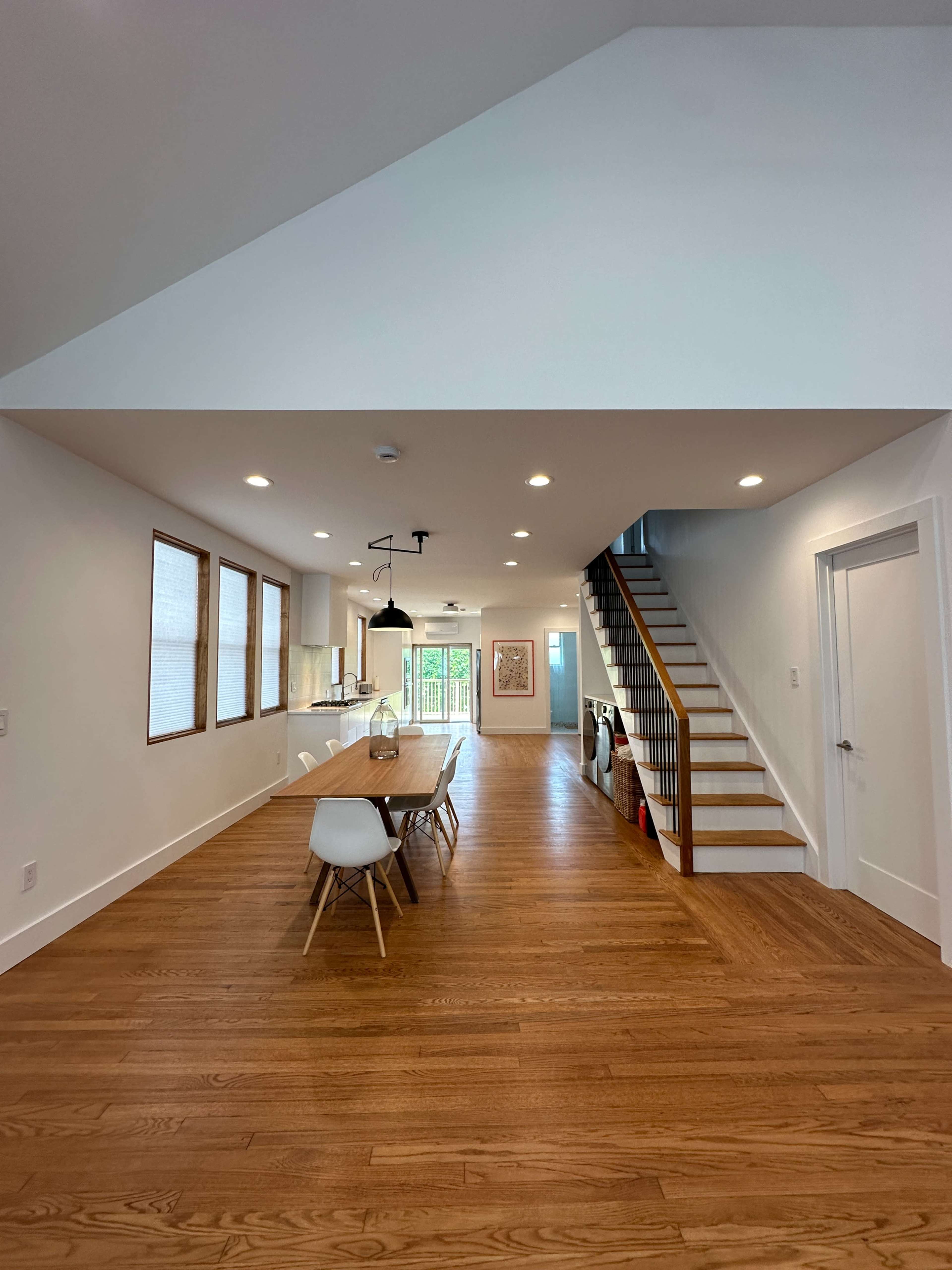 The image shows a modern open-concept living space featuring a dining area with a wooden table, a staircase on the right, and large windows providing natural light.