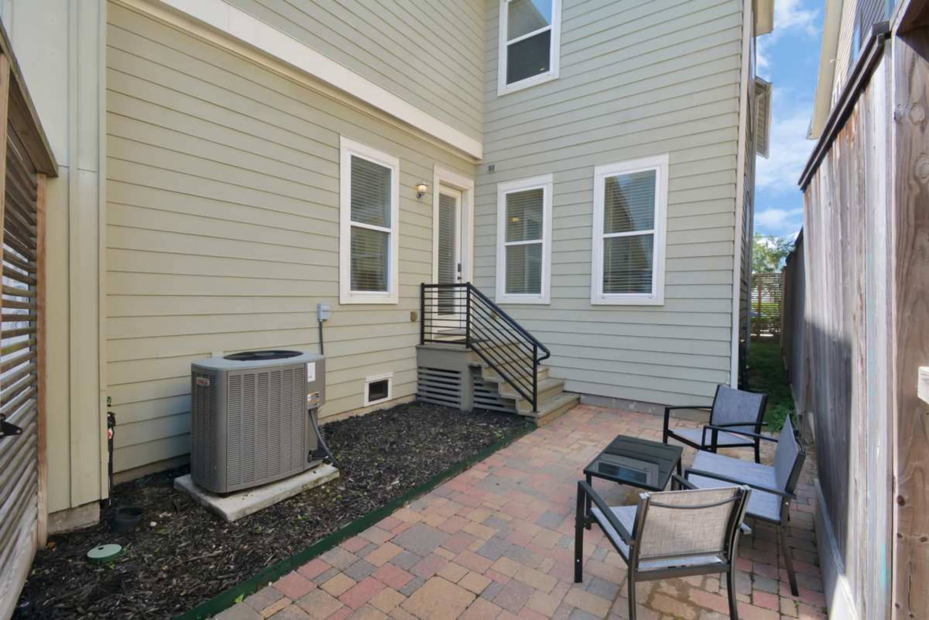 The image shows a small outdoor space with a paved patio featuring a few chairs and a small table next to the side of a two-story house, with an air conditioning unit visible.