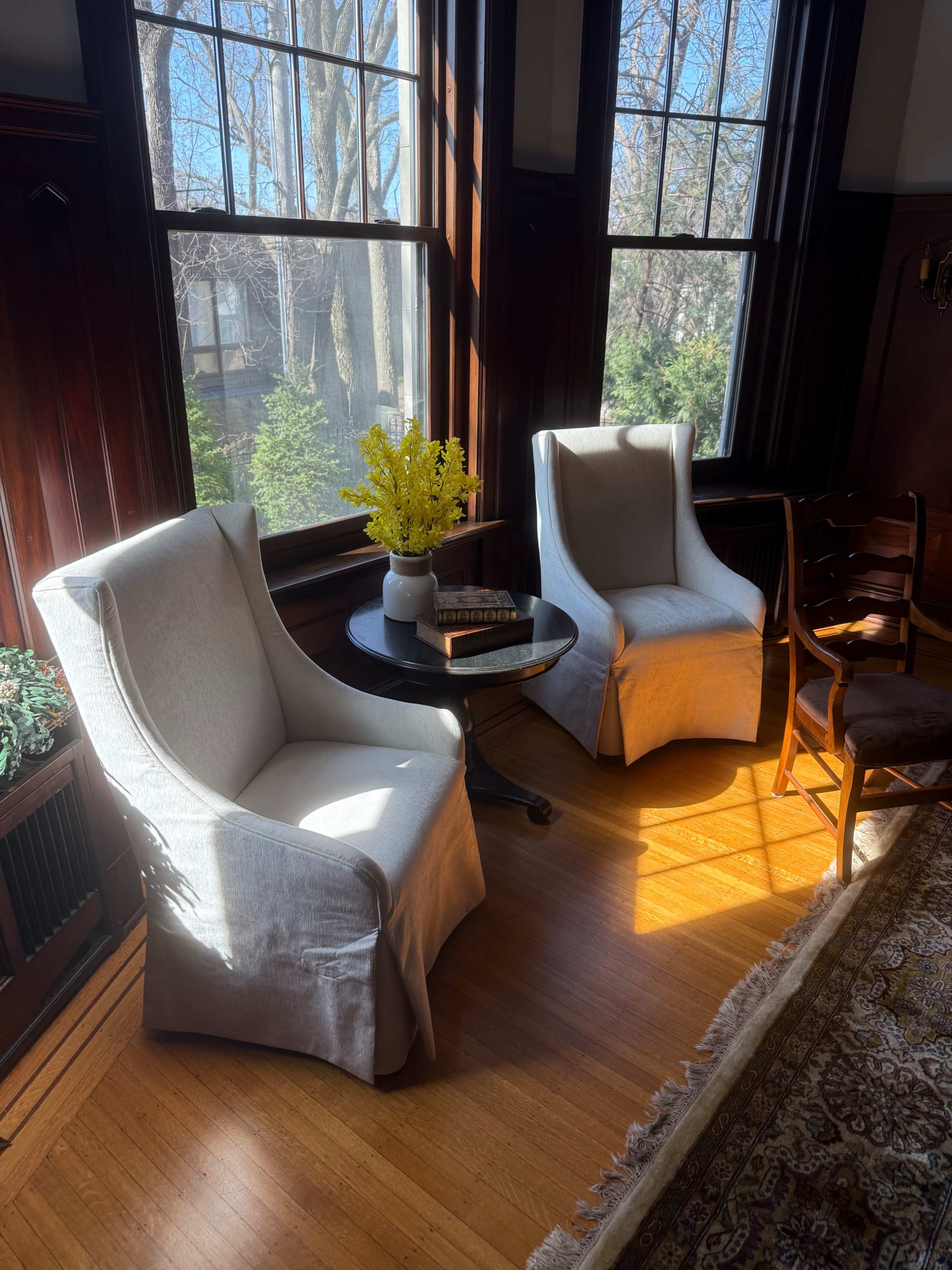Two chairs with light-colored fabric sit beside a small table, adorned with a vase of yellow flowers, under sunlight streaming through tall windows in a room with wooden accents.
