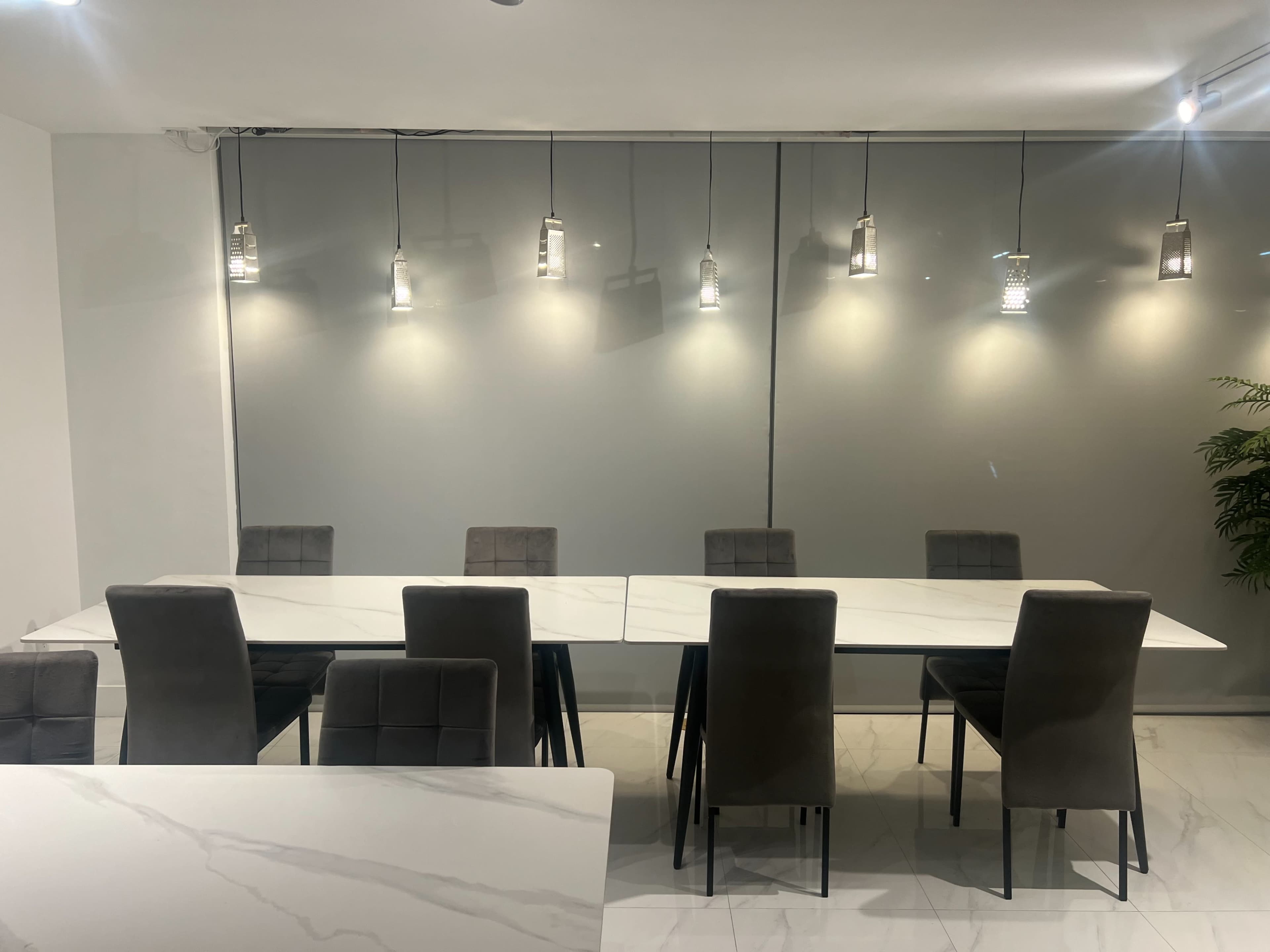 A modern dining area with a long marble table surrounded by several gray chairs and illuminated by pendant lights above.