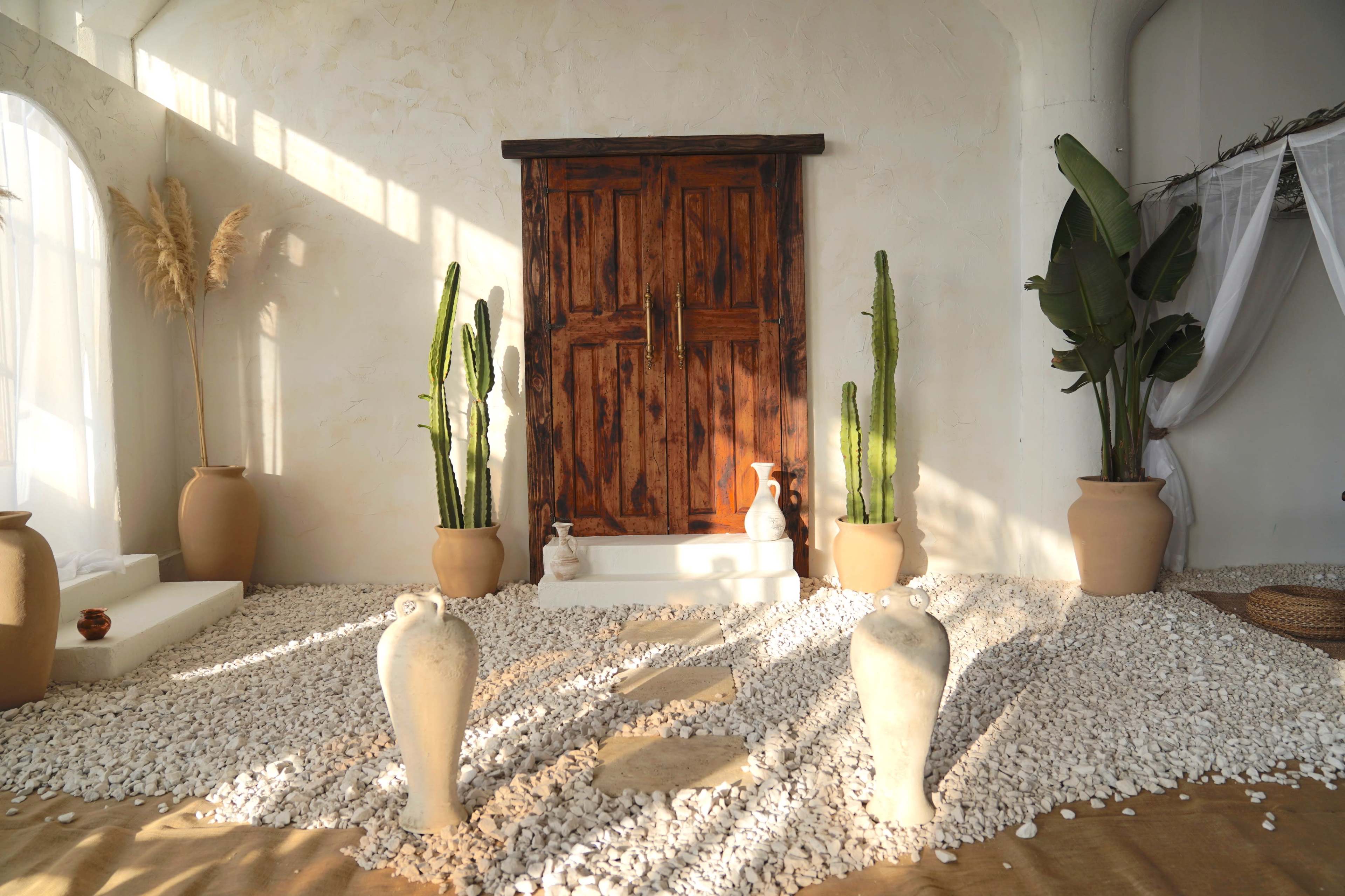 The image depicts a serene indoor space featuring a wooden door at the back, large potted plants, and a rocky, white pebble floor.