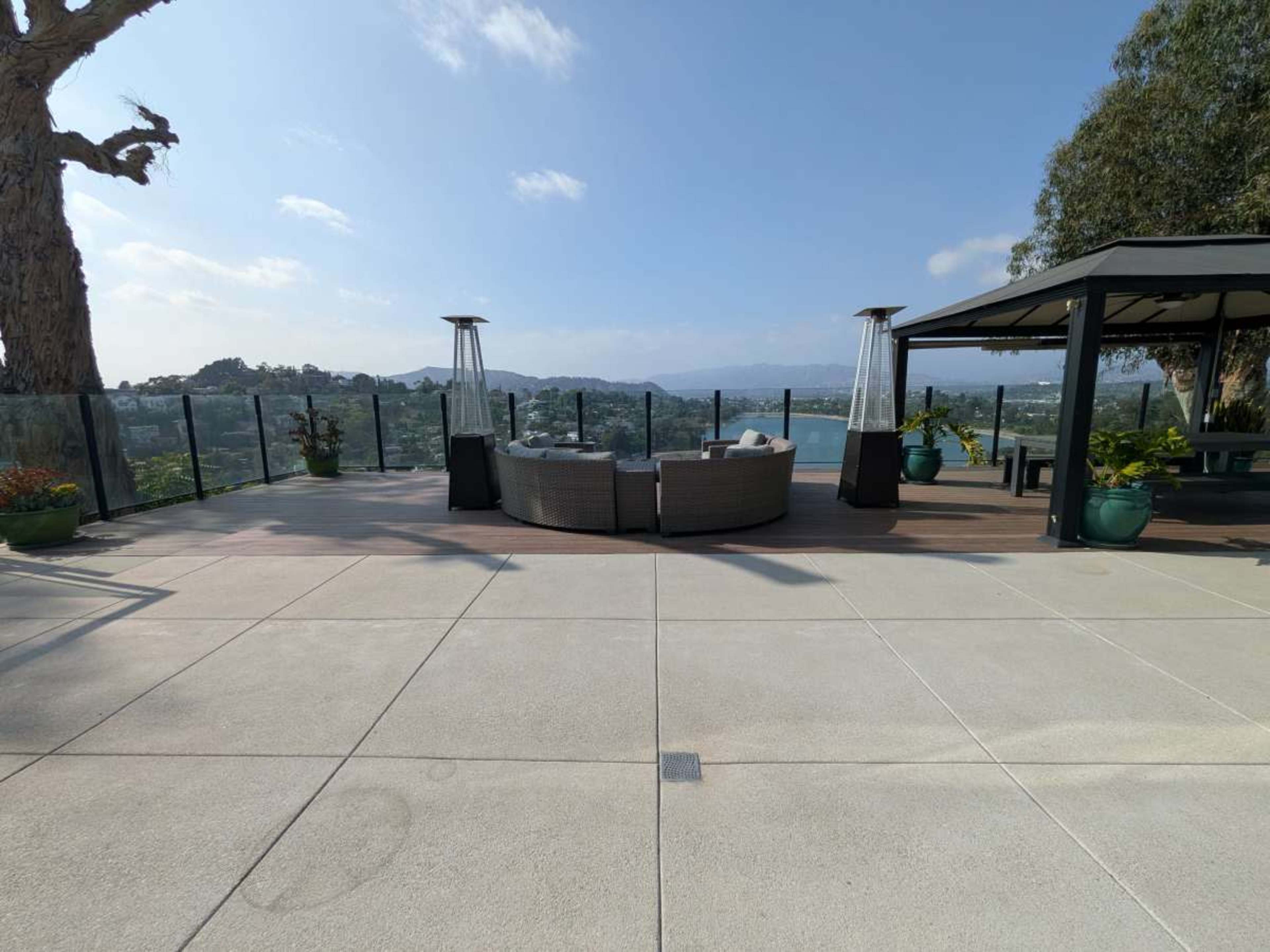 The image shows a spacious outdoor patio area with a circular seating arrangement, two outdoor heaters, and a view of surrounding hills and water.