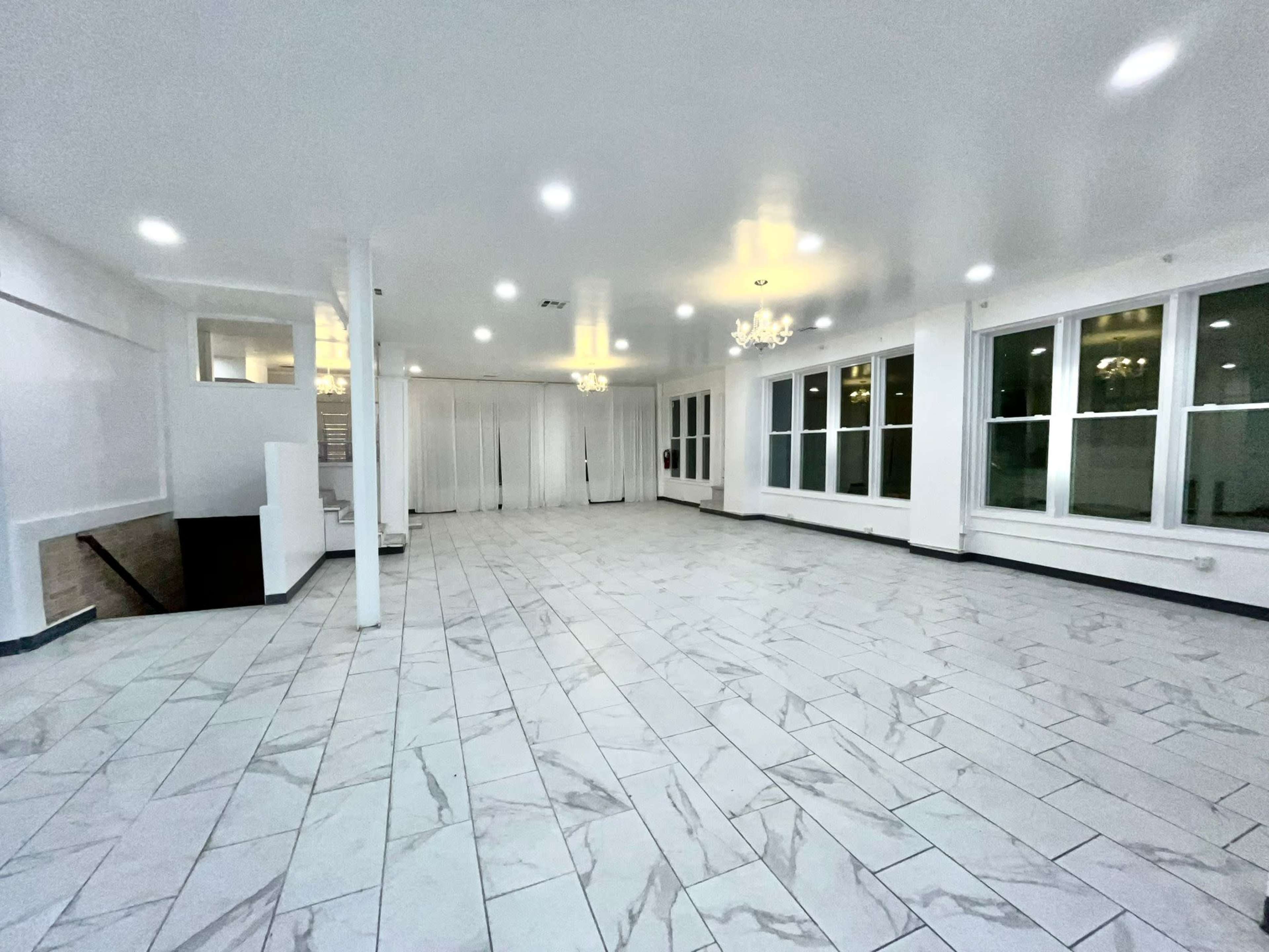 A spacious, well-lit room features large windows, white walls, and a tiled floor with a chandelier overhead.
