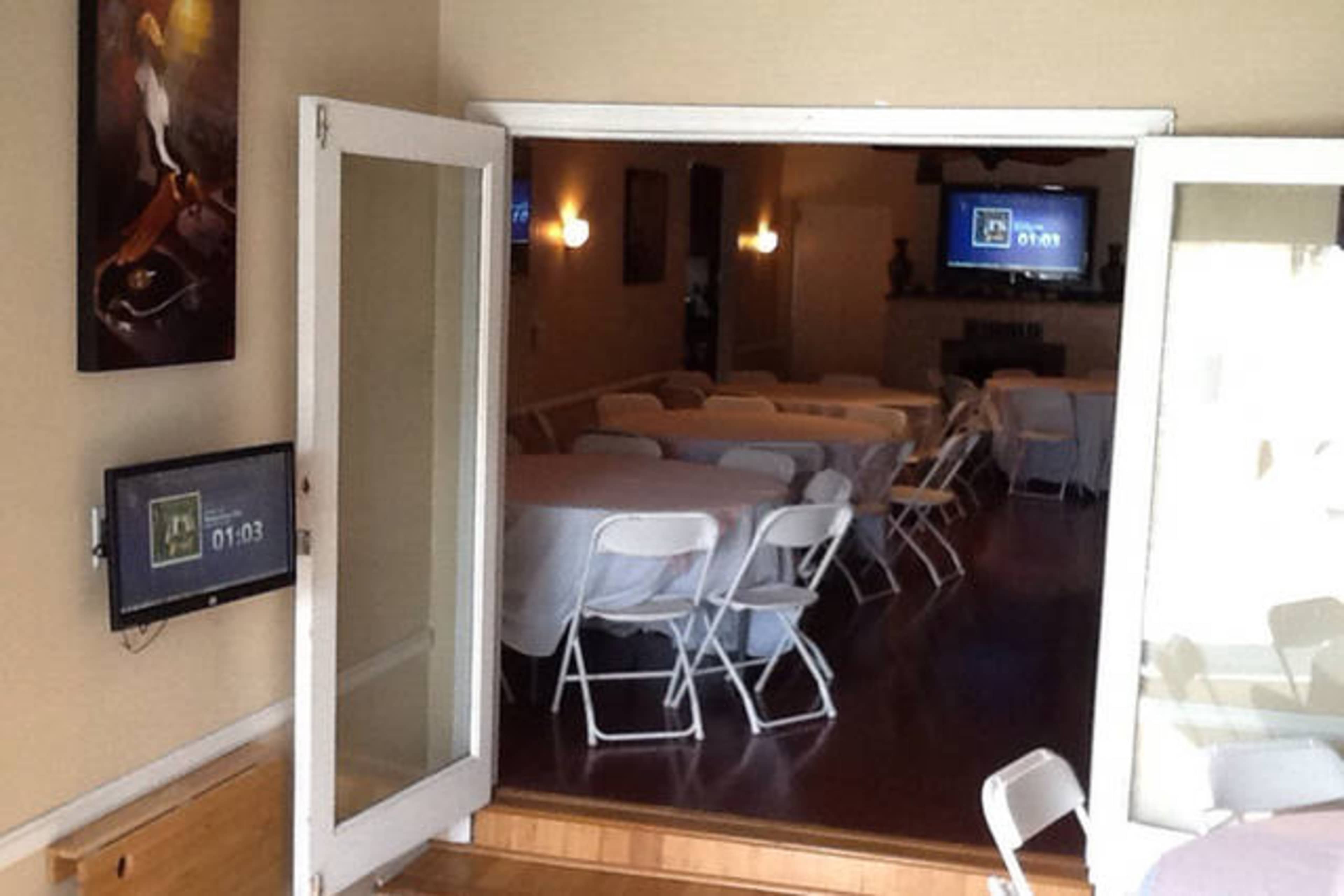 An interior room with several tables covered in white tablecloths and arranged with plastic chairs, visible through open double doors.