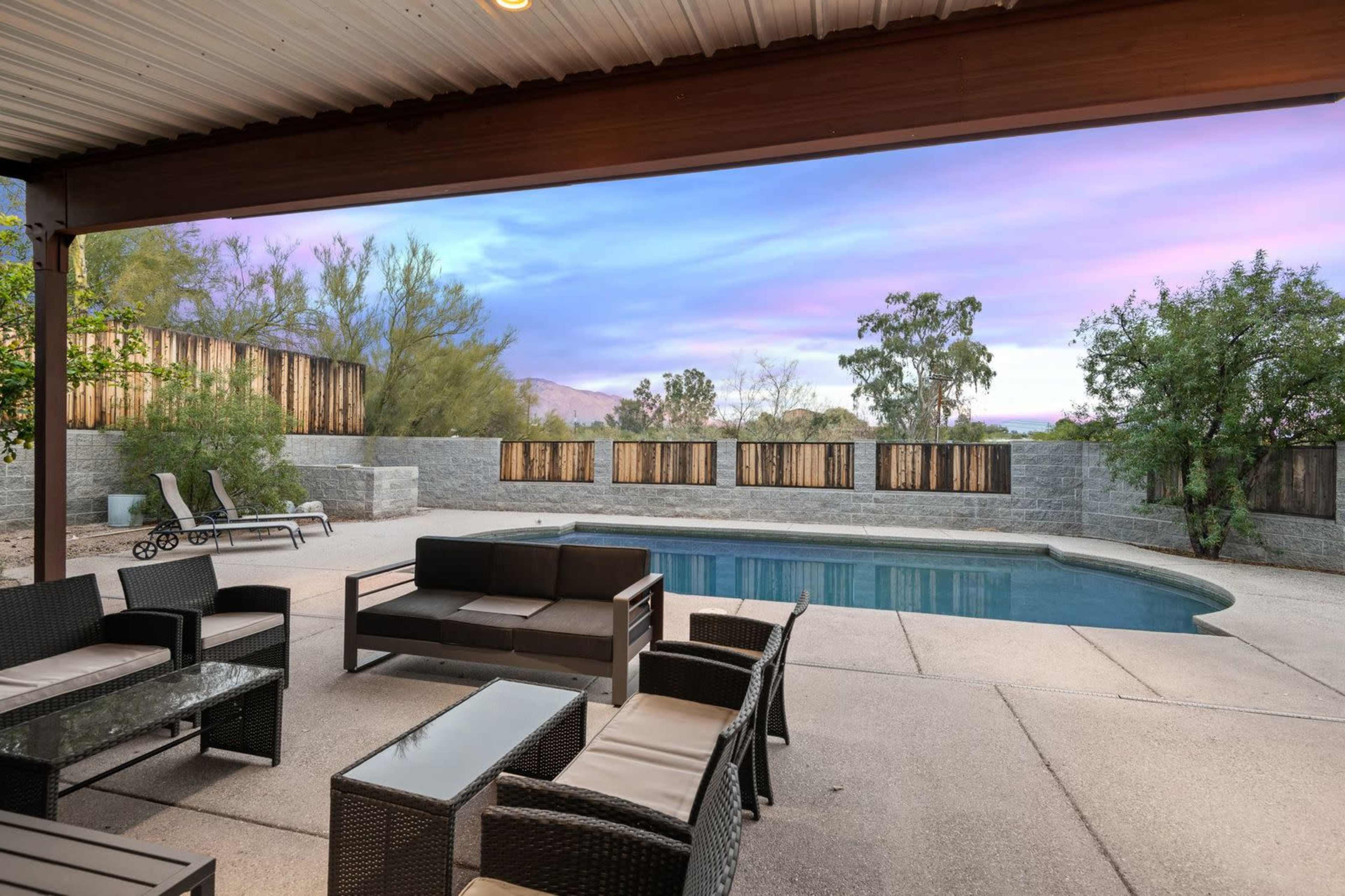 The image shows a backyard with a swimming pool, outdoor seating, and a fence surrounded by greenery under a colorful sunset sky.