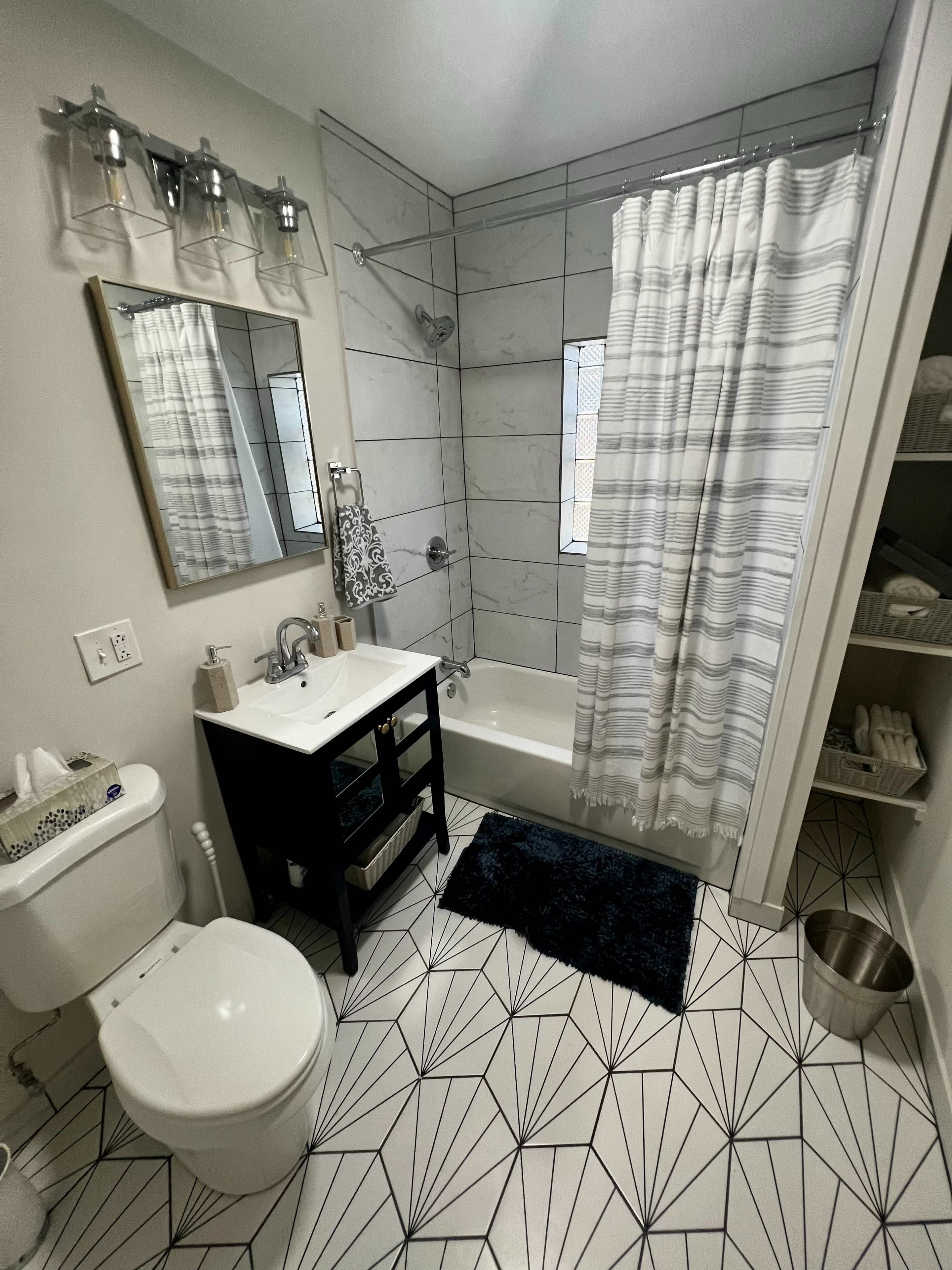 The image shows a modern bathroom featuring a bathtub with a shower curtain, a vanity sink with a mirror, a toilet, and patterned tile flooring.