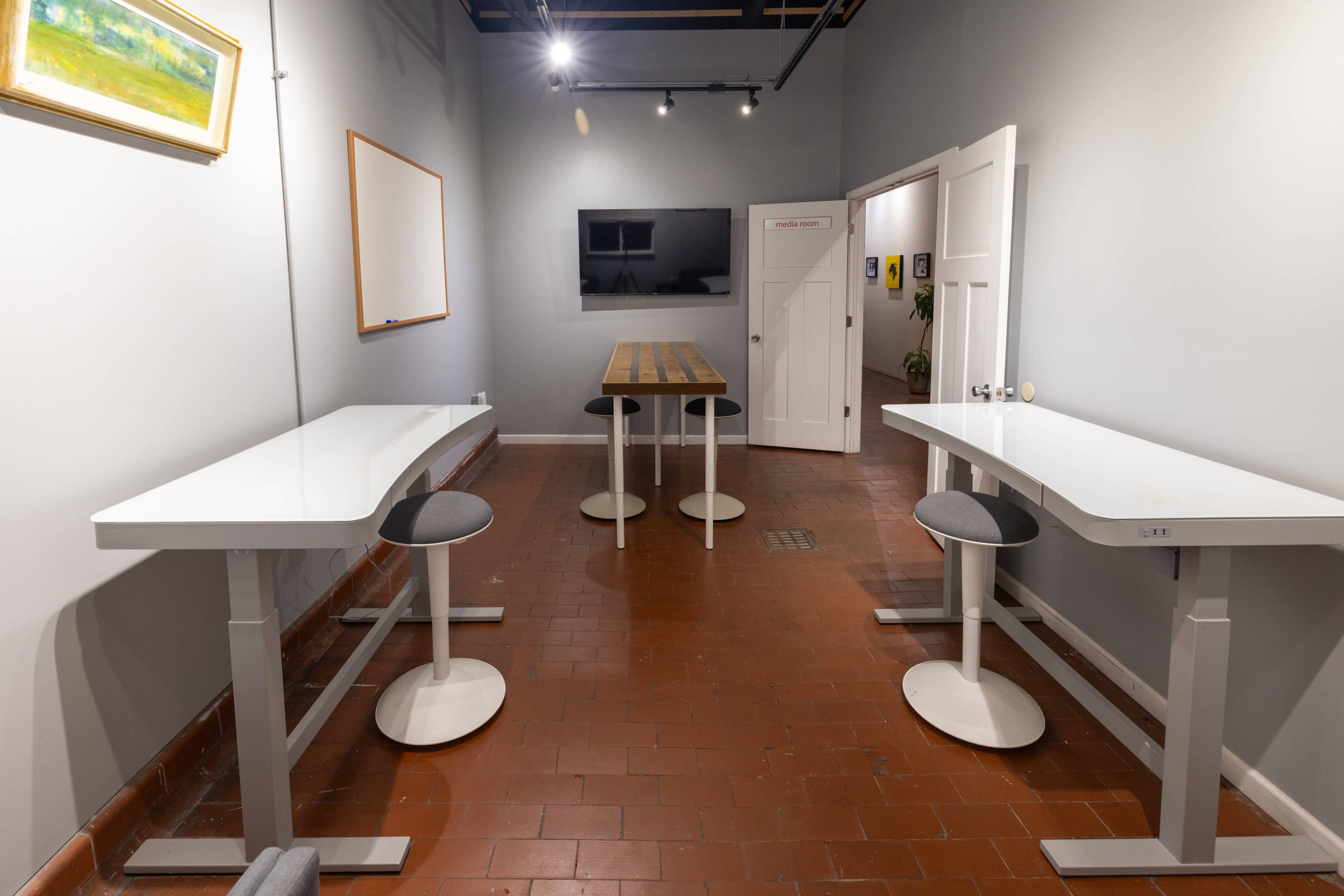 A spacious room with two high tables on one side, a long wooden table in the center, and a wall-mounted television opposite a whiteboard.