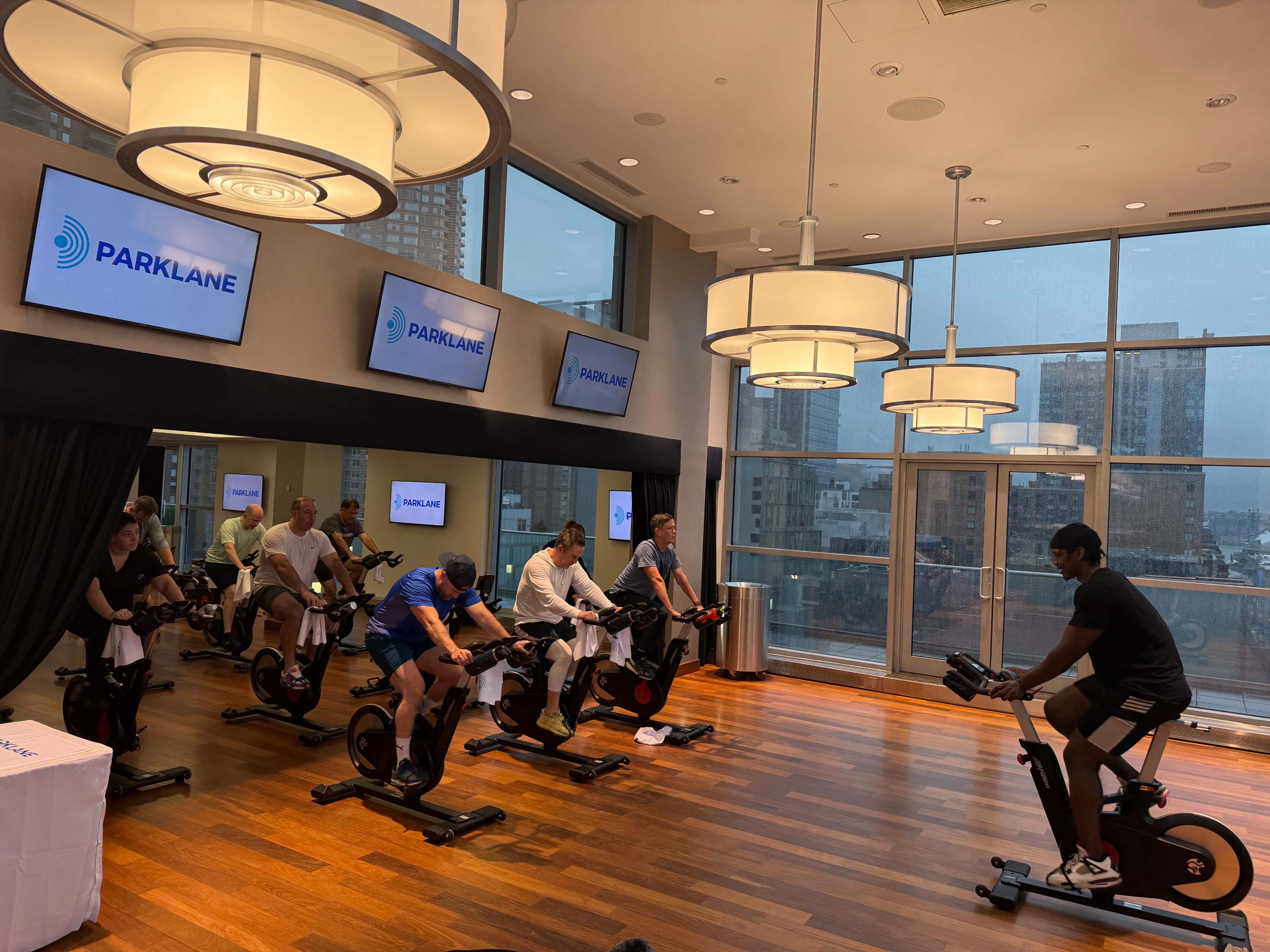 A group of people are participating in a spinning class indoors, with large windows displaying a cityscape in the background.