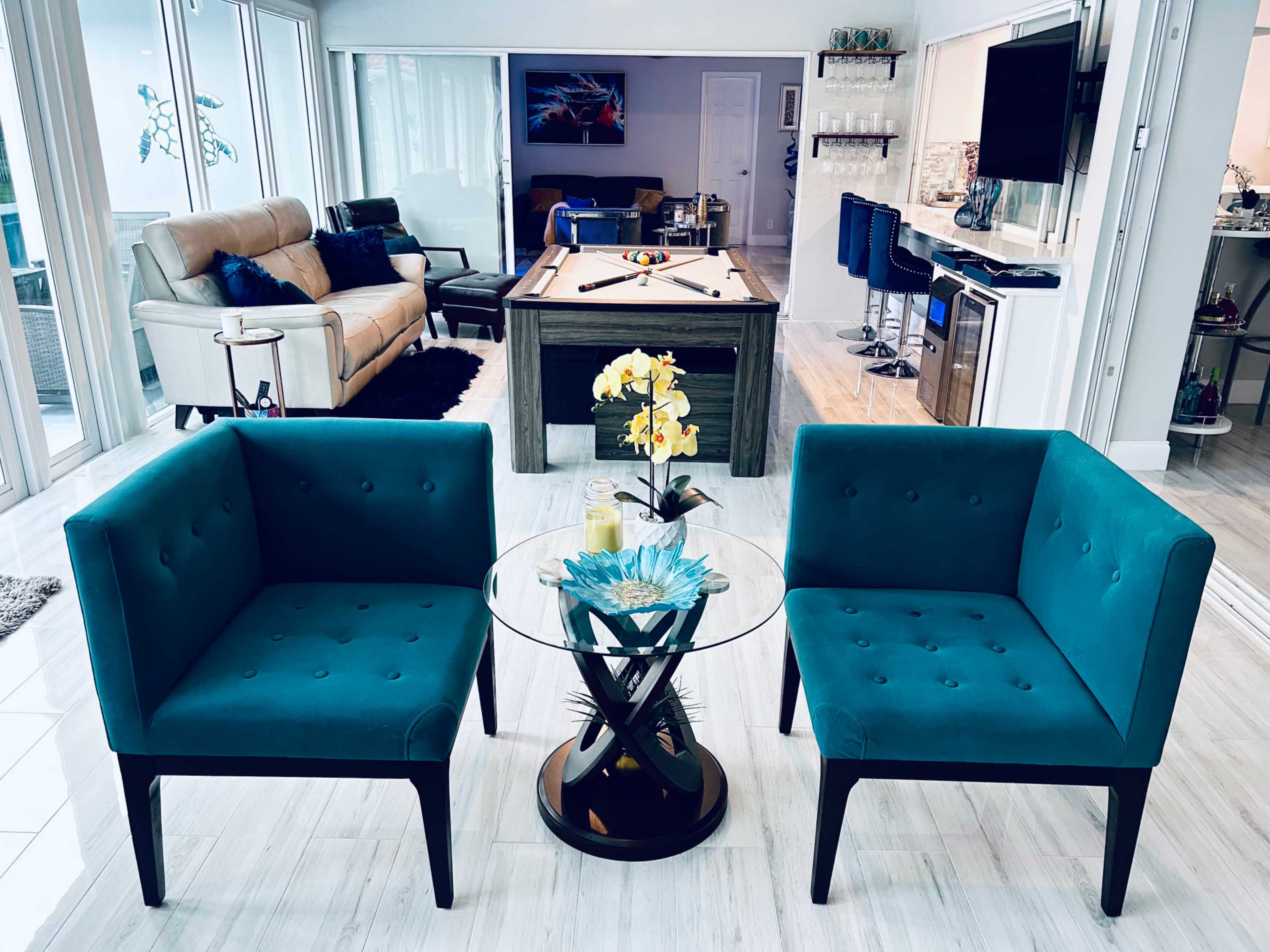 A modern living space with two turquoise chairs, a glass coffee table, and a view of a pool table and a seating area in the background.