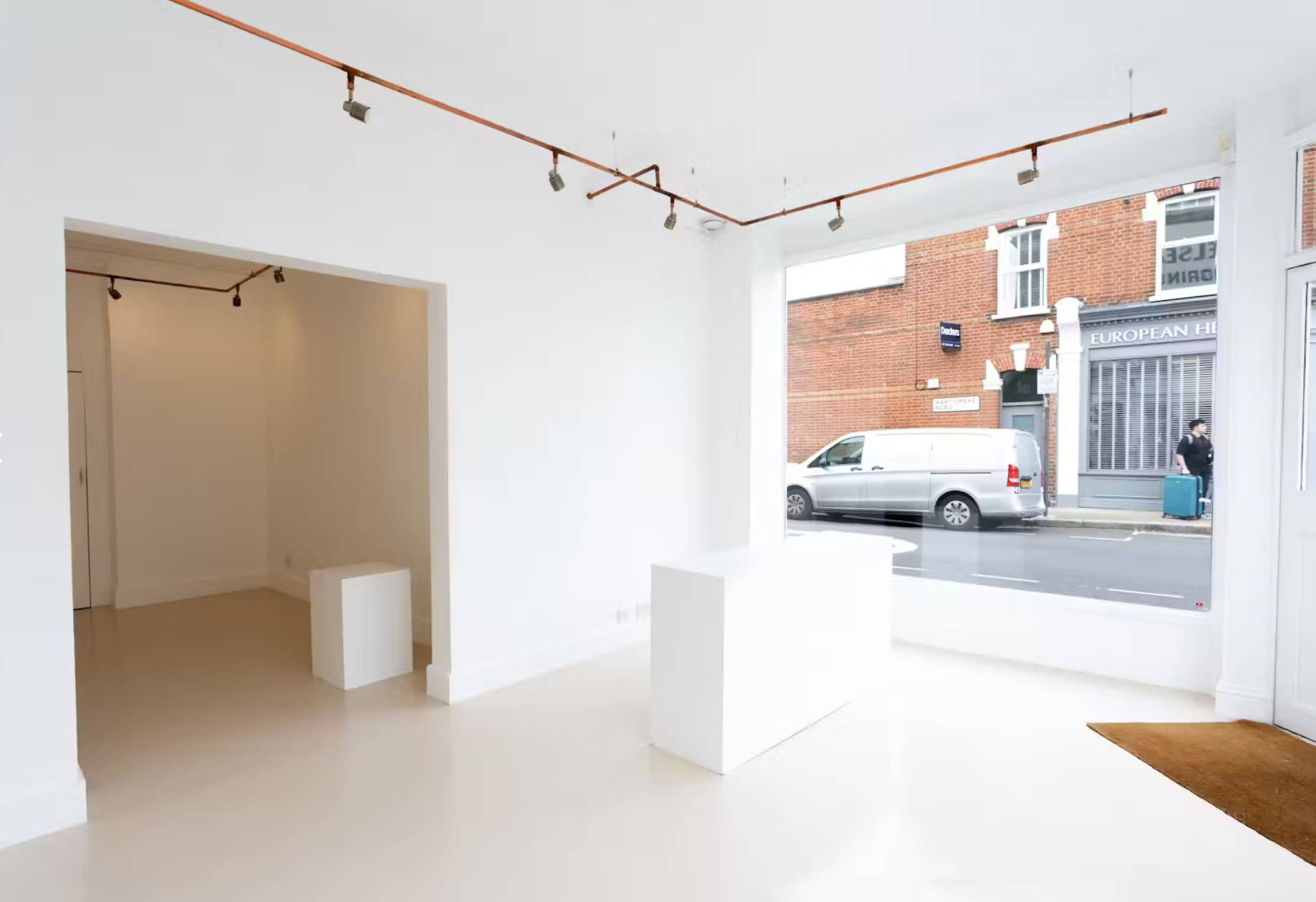The image shows a minimalist, white-walled interior space featuring a small podium and a view of a street through large windows.