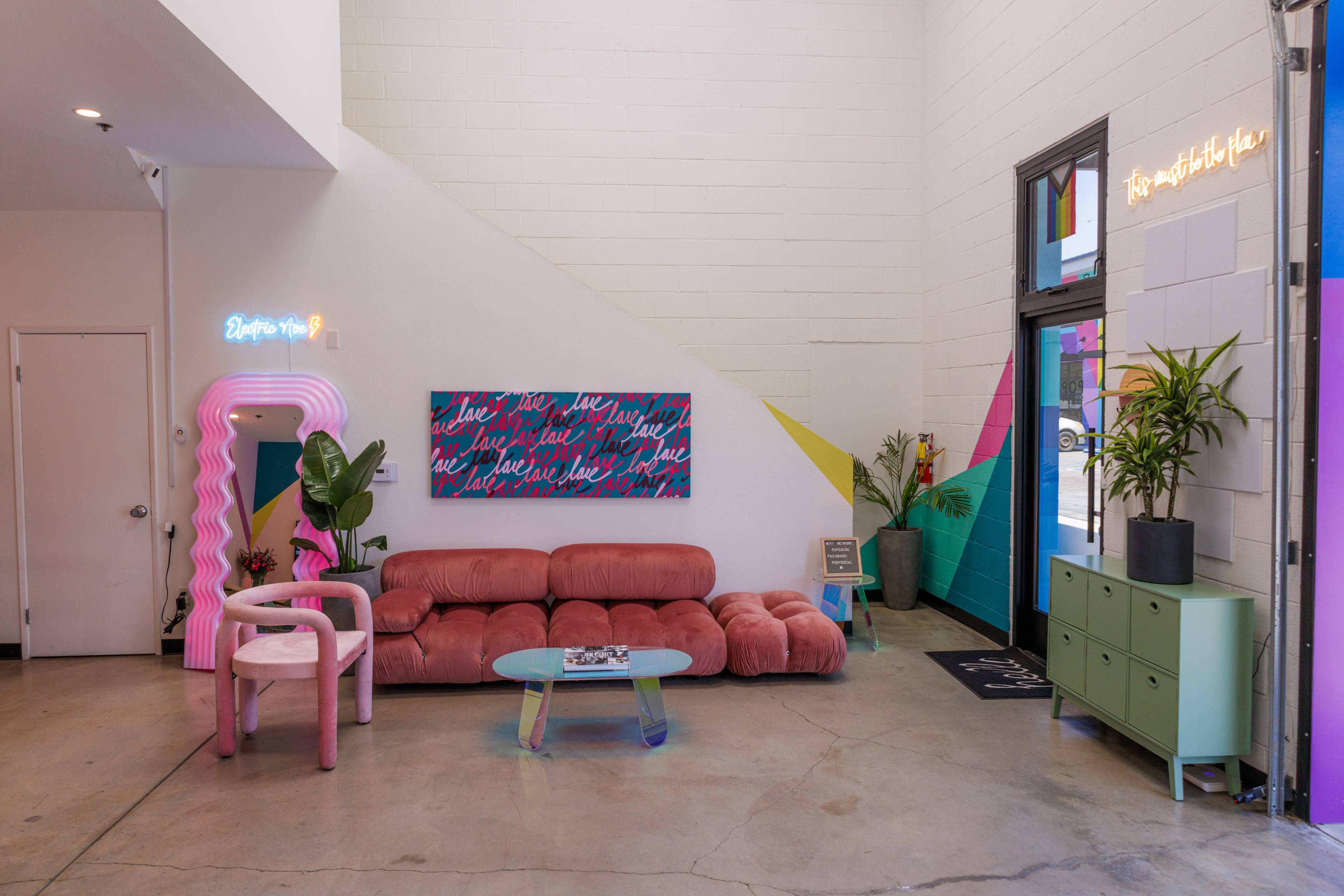 The image shows a modern interior space with a pink couch, a green cabinet, a wall-mounted artwork, plants, and neon signs, featuring a mix of geometric shapes and bright colors.