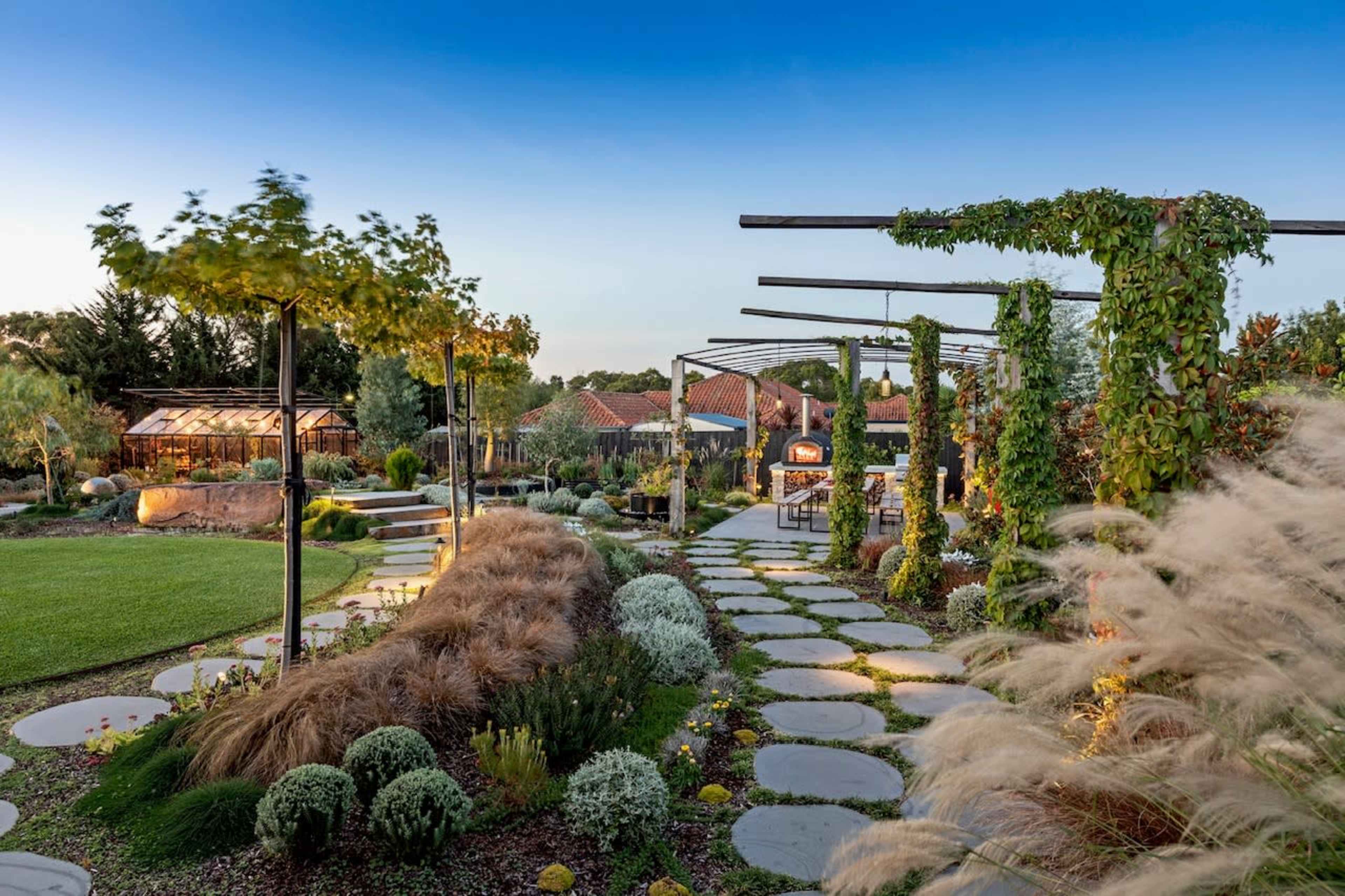 A landscaped garden features stone pathways, greenery, and a covered seating area under a blue sky.