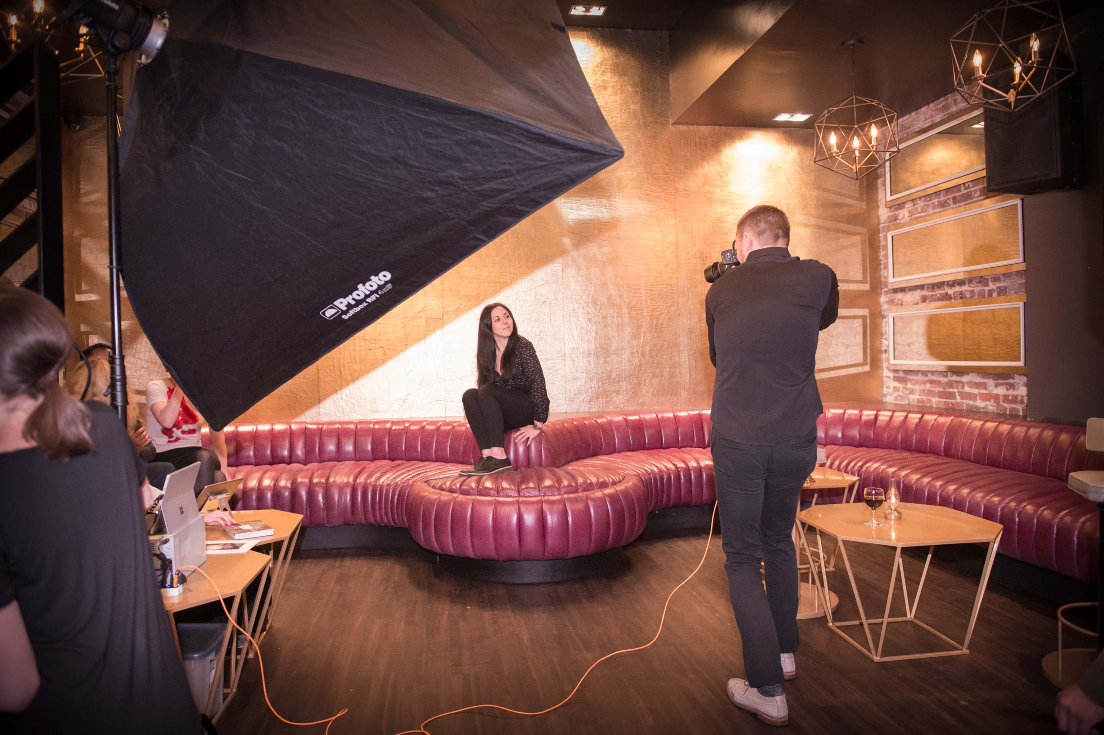 A photographer captures a model sitting on a curved, upholstered seating area in a stylish, dimly lit lounge.