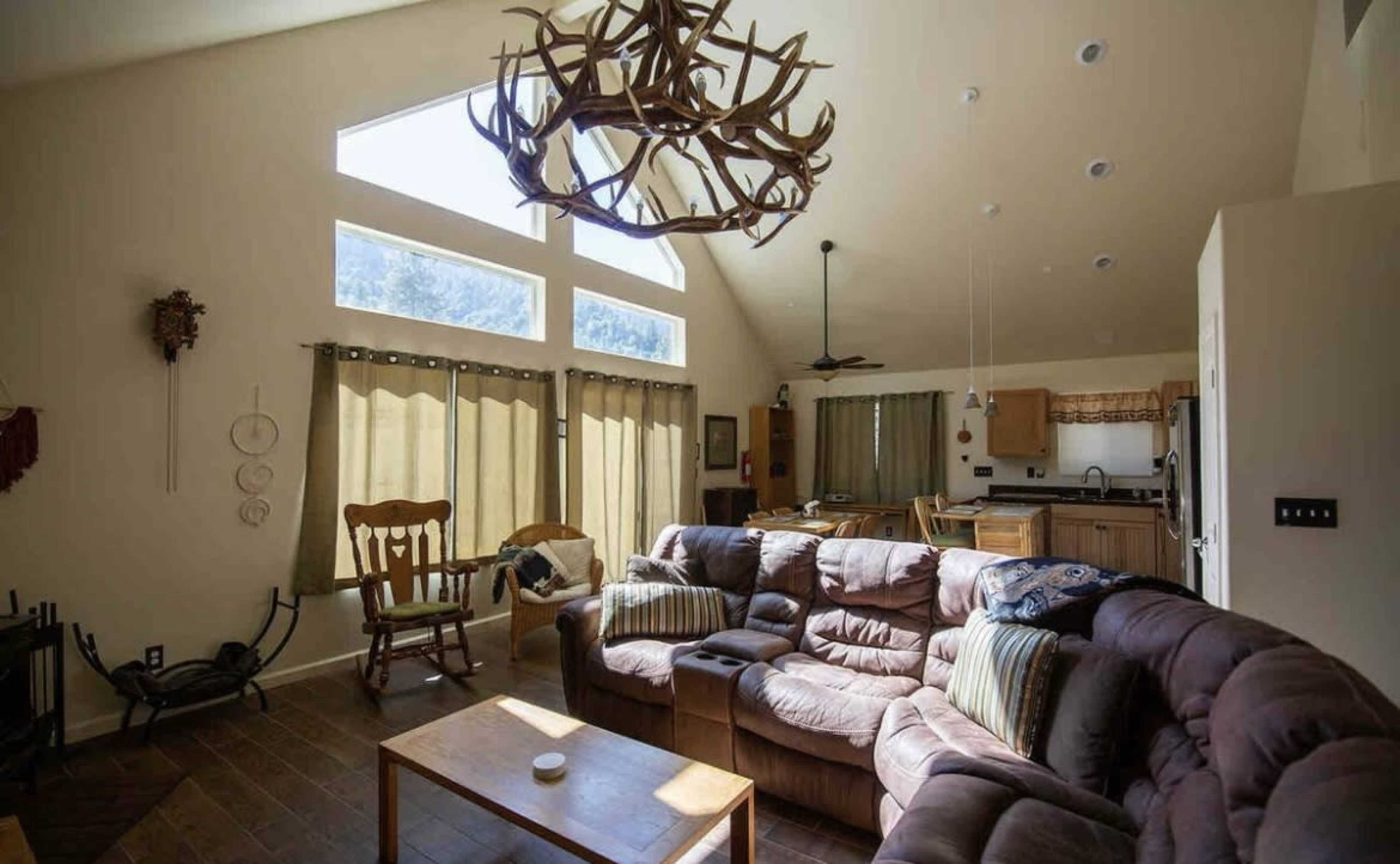 A spacious living room with large windows, a brown sectional sofa, and a decorative antler chandelier.