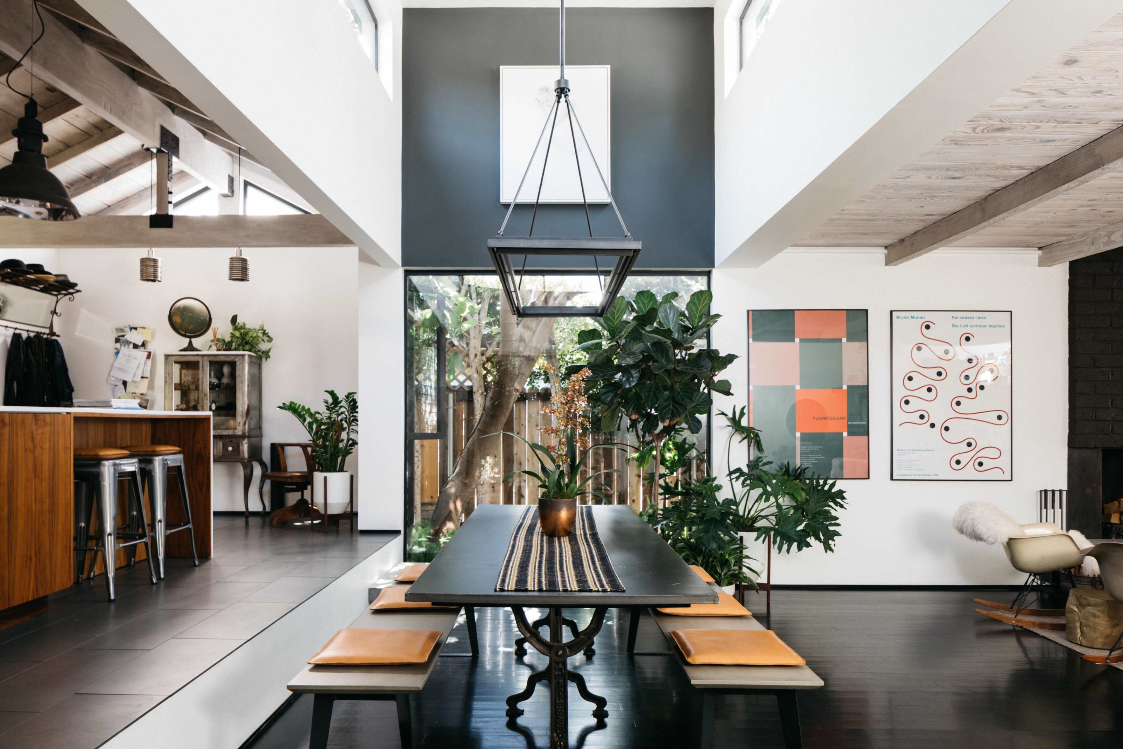 A modern dining area with a dark wooden table surrounded by benches, large plants, and artwork, illuminated by a geometric pendant light.