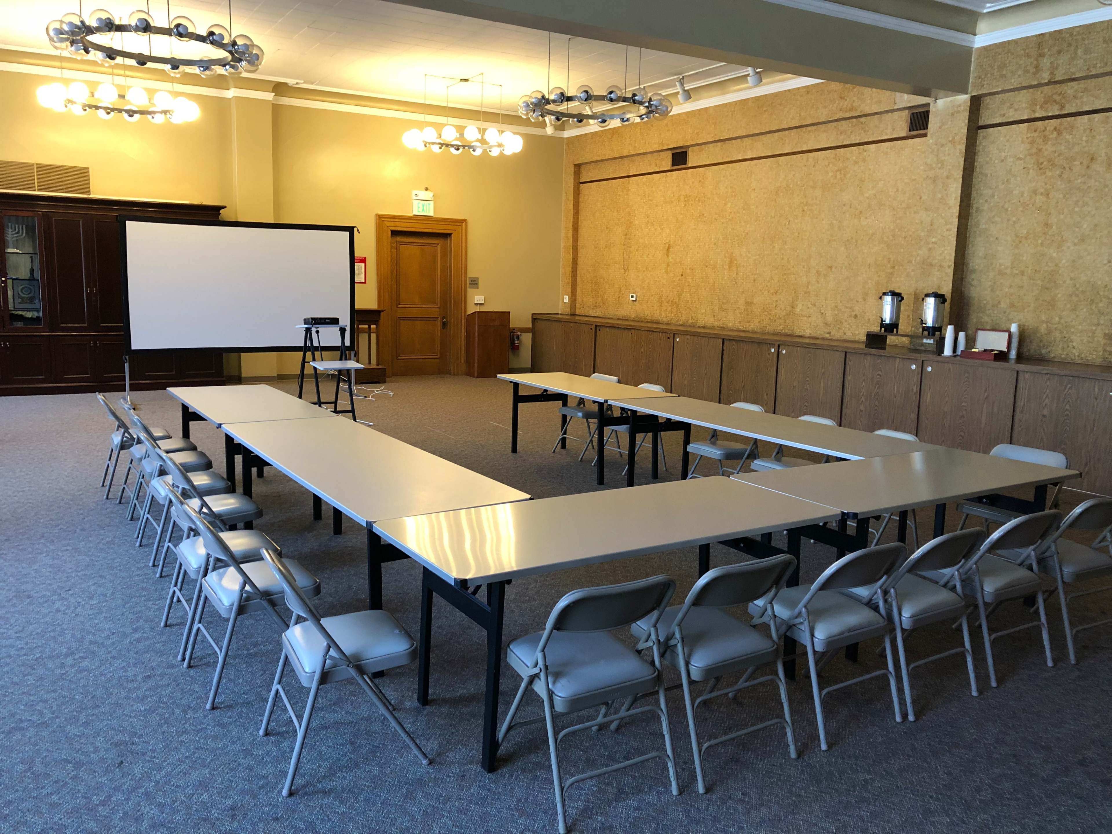 A meeting room features a rectangular arrangement of tables and chairs, with a projection screen at the front and coffee service on a side counter.