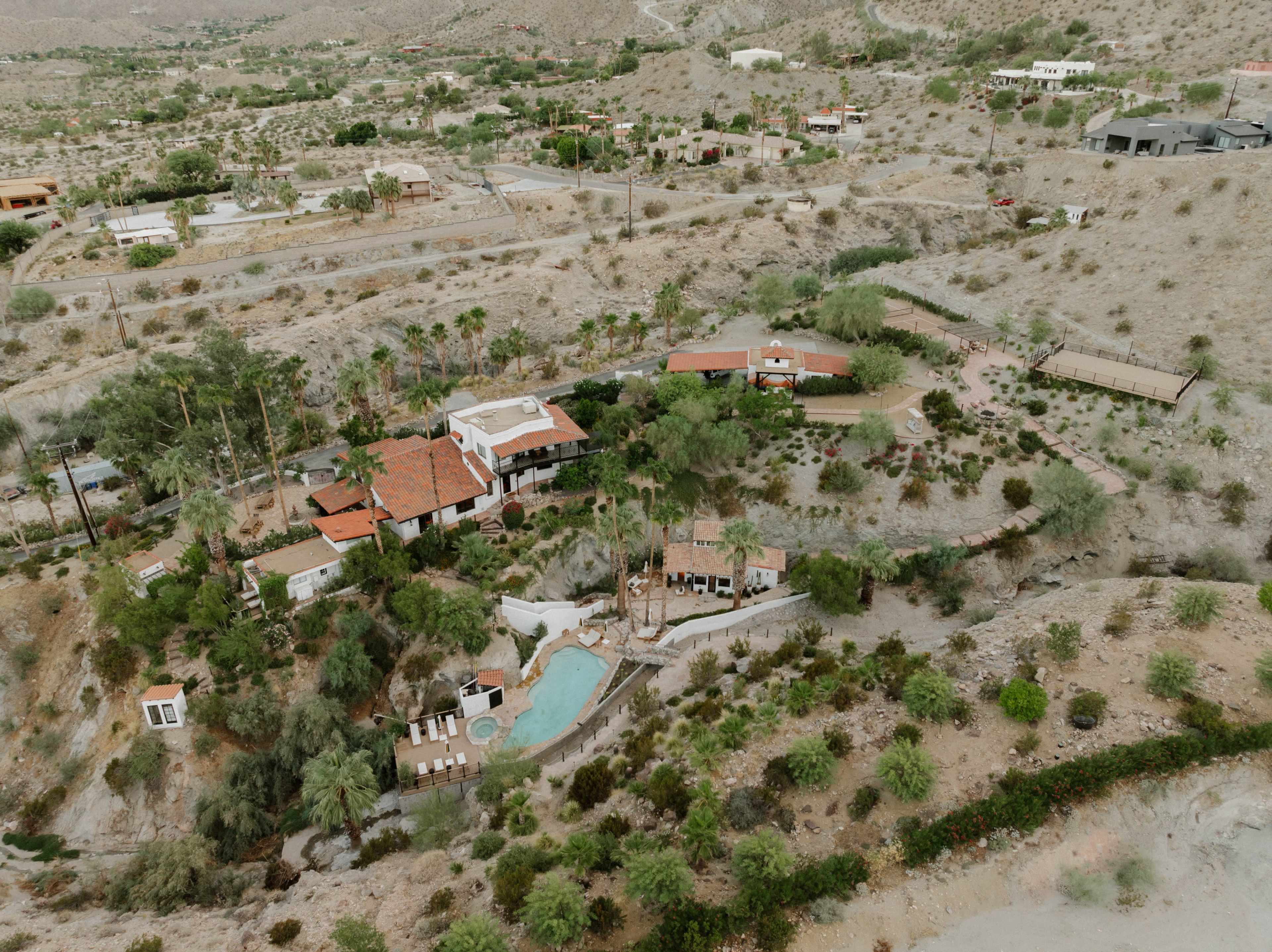 The image shows an aerial view of a desert landscape featuring a residential property with a swimming pool, surrounded by palm trees and sparse vegetation.