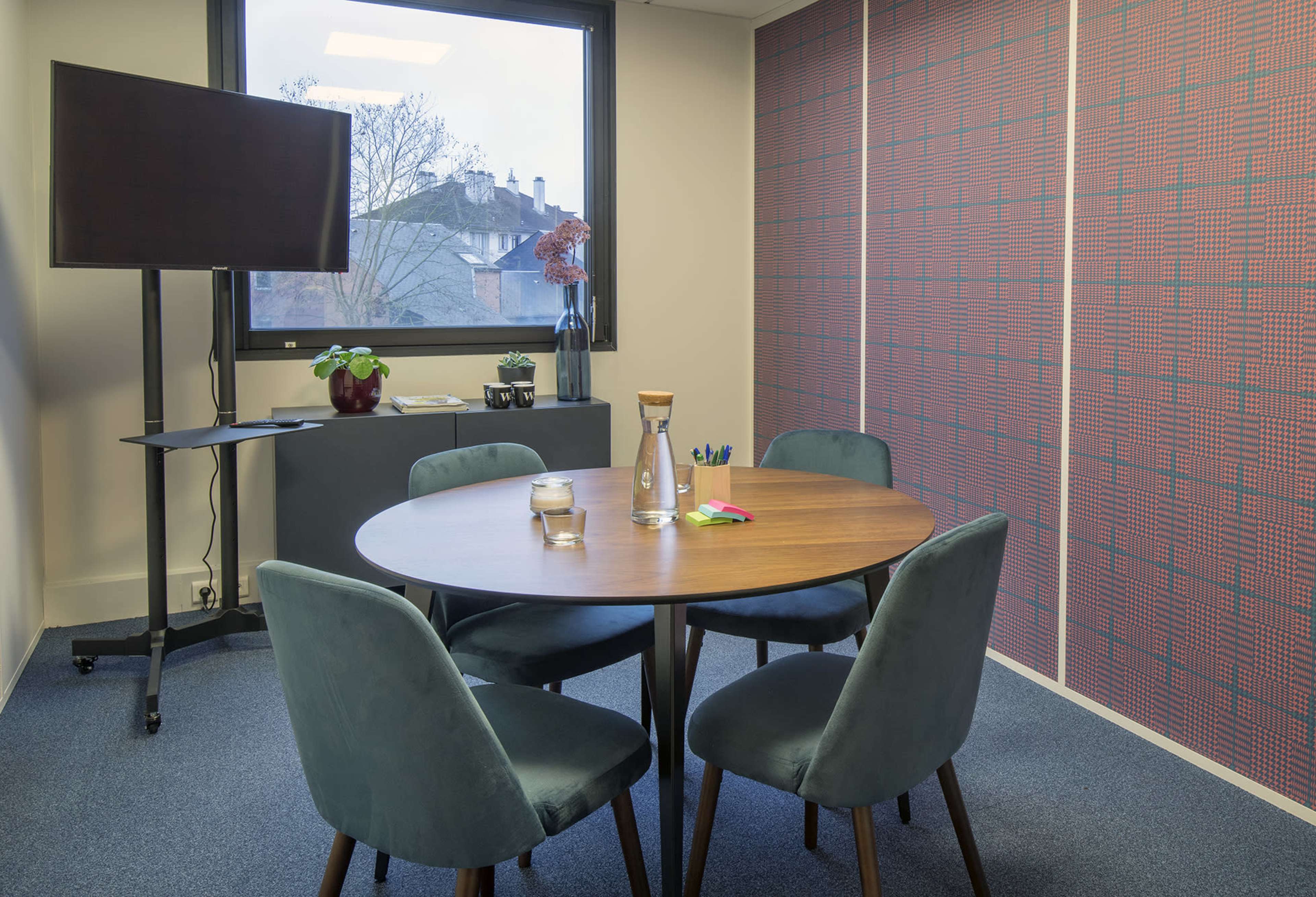 A round wooden table is surrounded by four green upholstered chairs in a small meeting room with a television on a stand and a patterned wall.