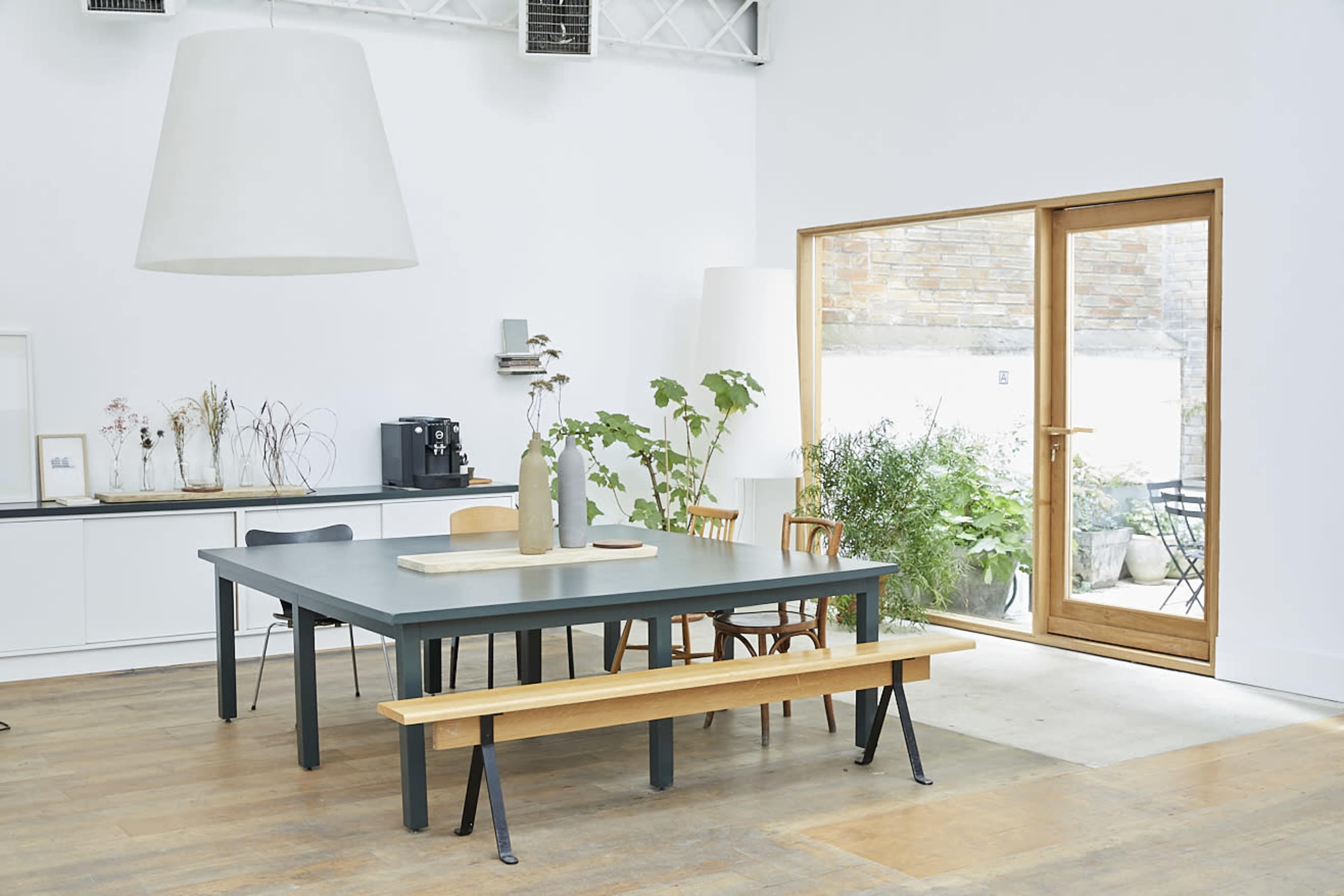 A modern dining area features a large table with wooden chairs and a bench, surrounded by bright plants and large windows.