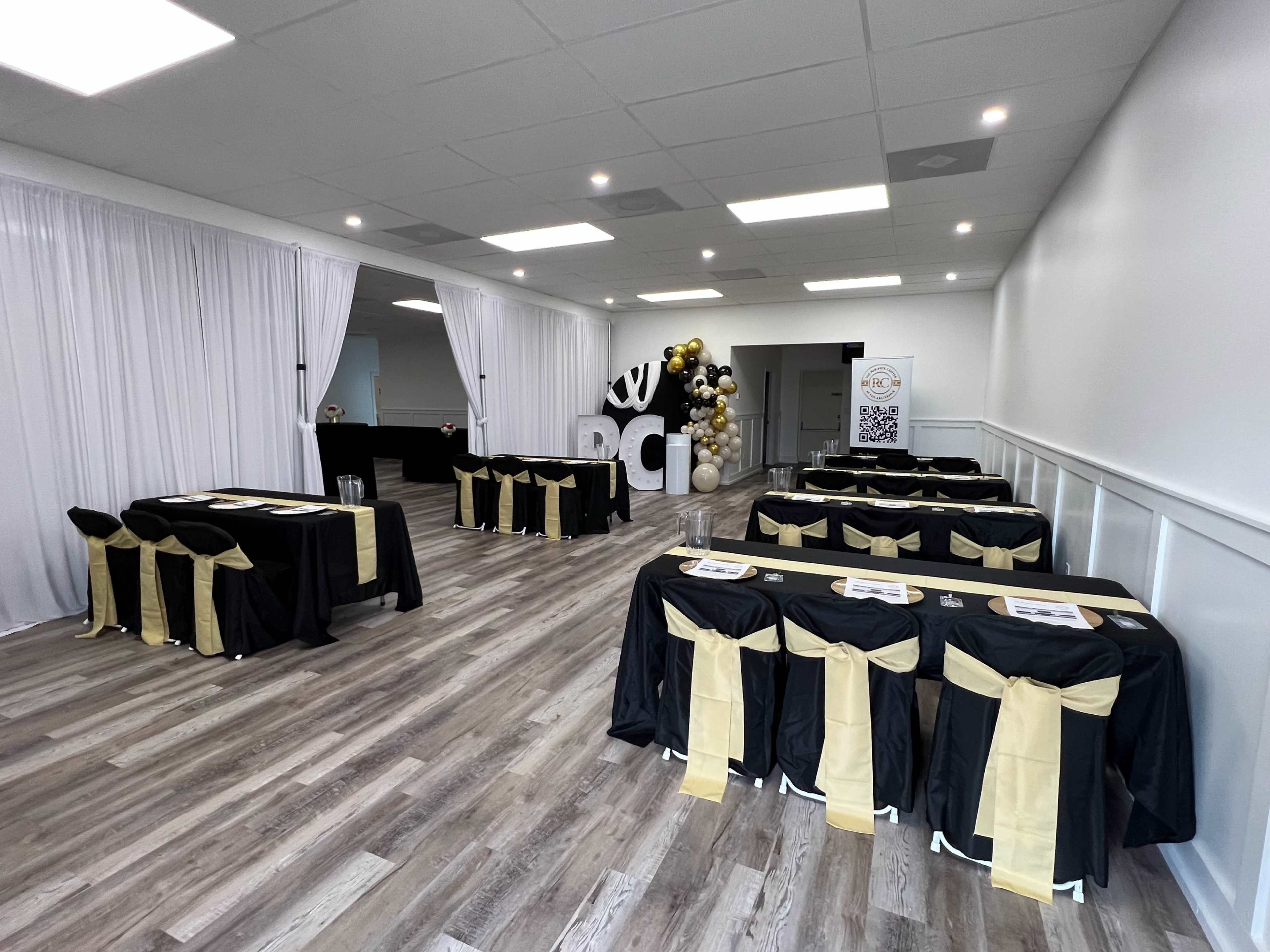 A spacious event room set up with several tables covered in black cloth with gold accents, surrounded by white drapery and a decorative balloon arch.