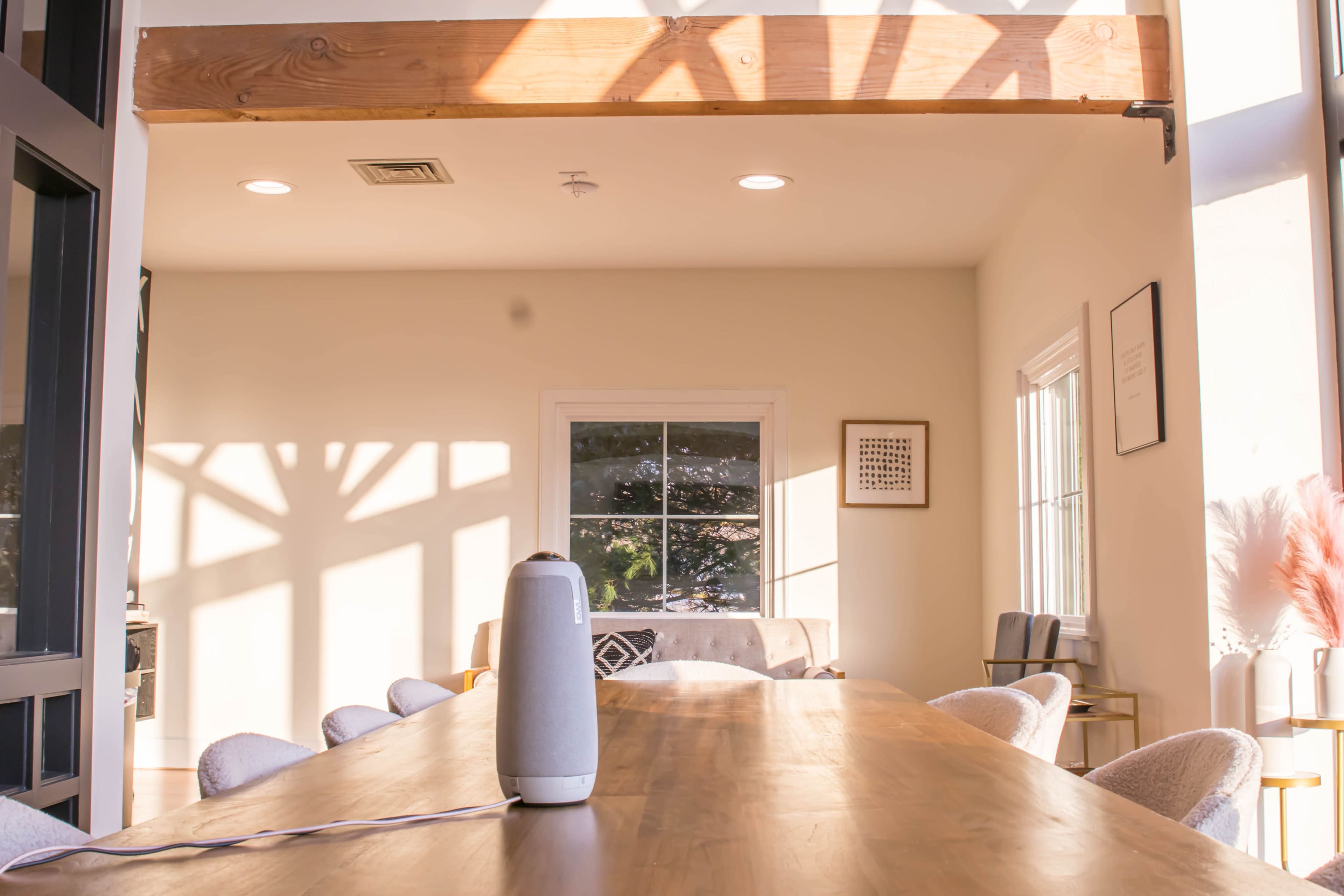 A modern dining area features a long wooden table with a speaker on it, illuminated by natural light streaming through a window.