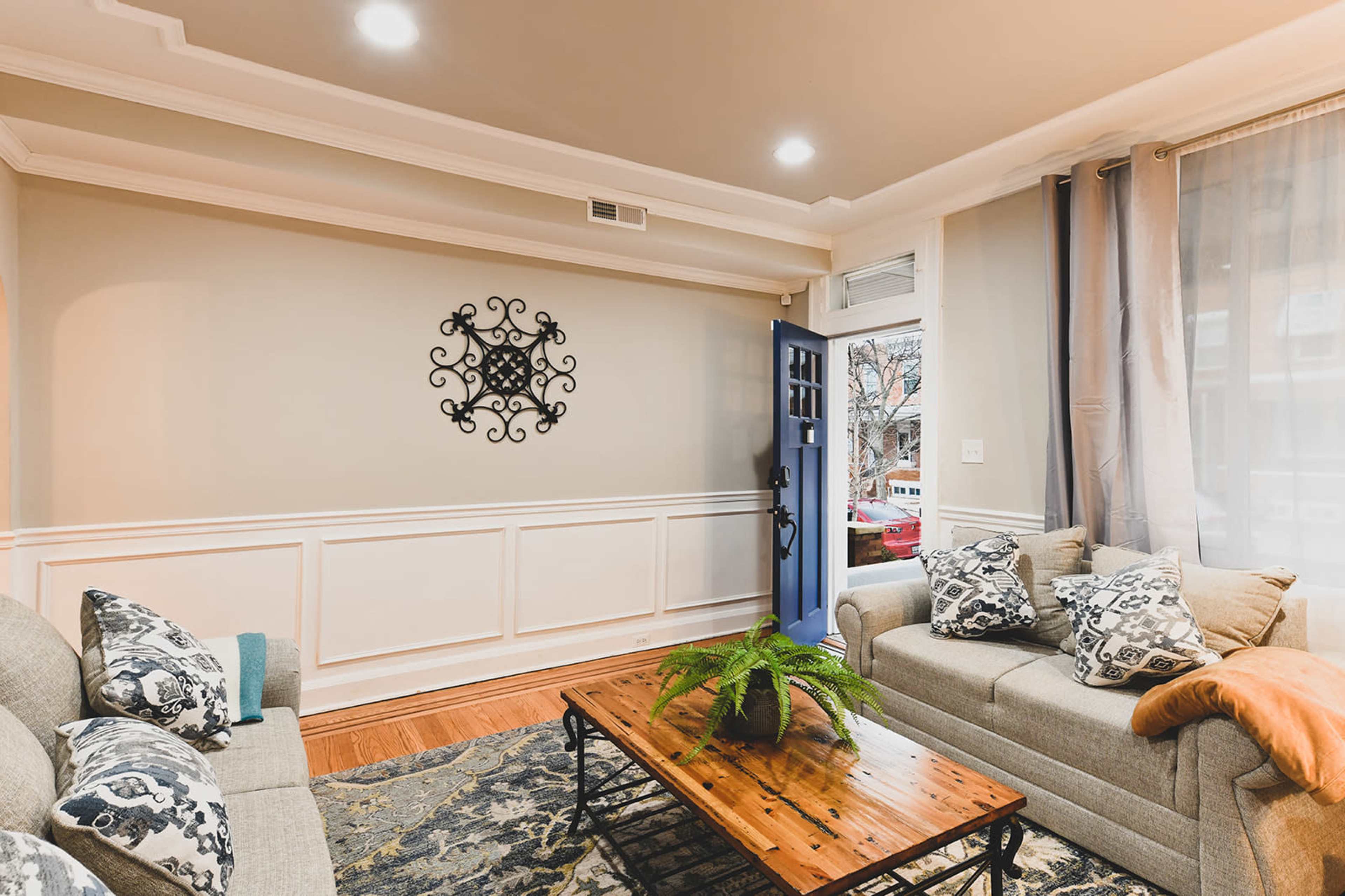A living room features two sofas, a wooden coffee table with a plant, and a blue front door opening to the outside.