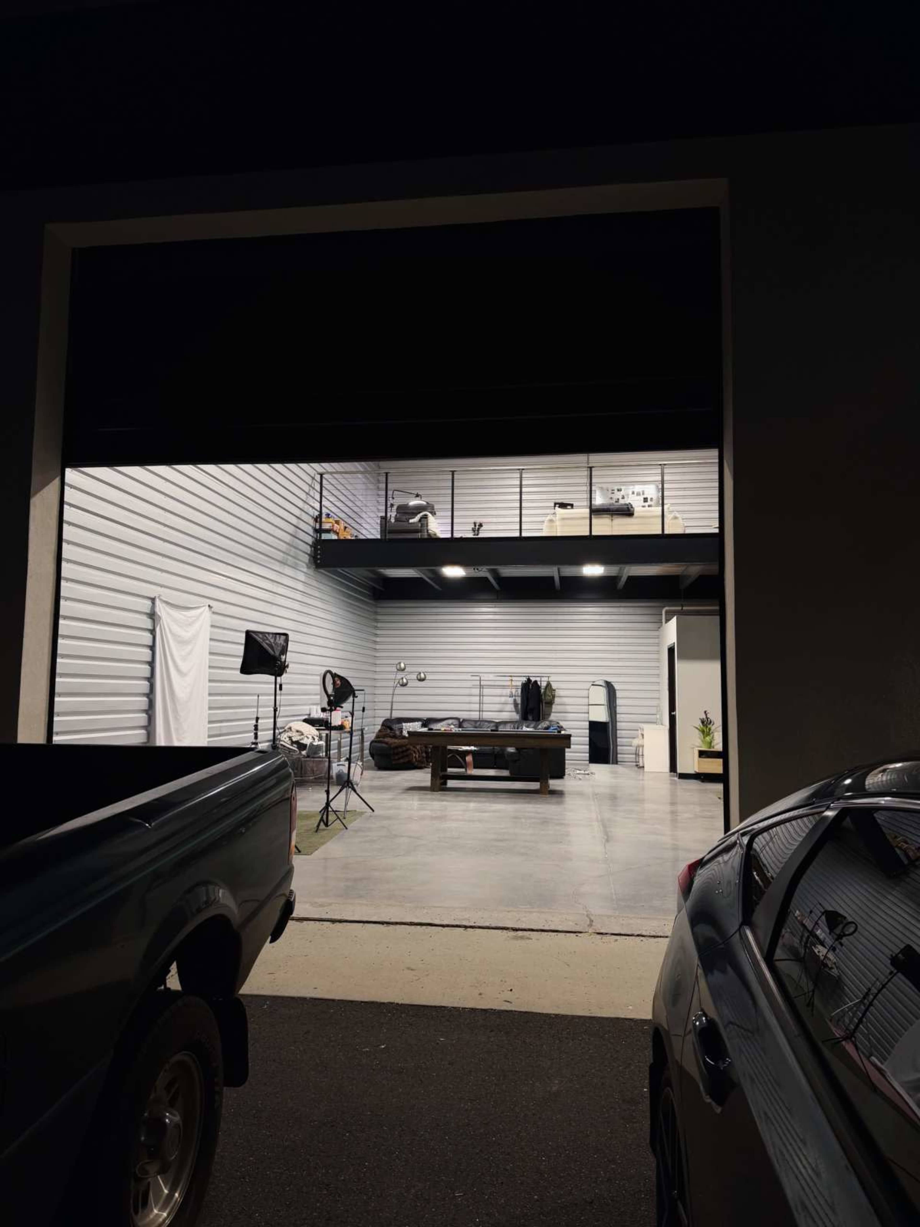 A spacious garage features a loft area with various items, including a bed and furniture, illuminated by overhead lights.
