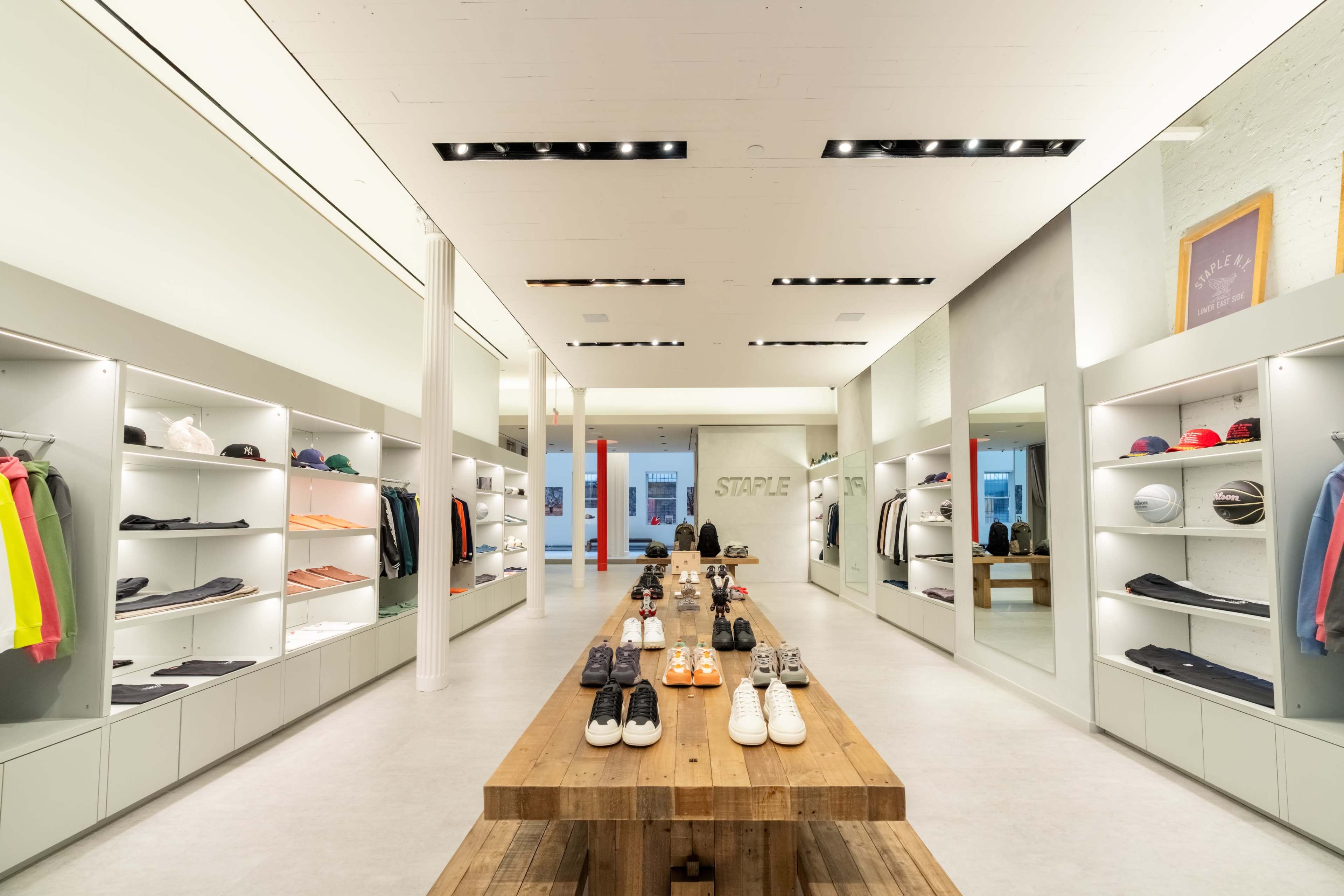 The image shows a modern retail store interior featuring neatly arranged apparel and footwear on display shelves and a central wooden table.