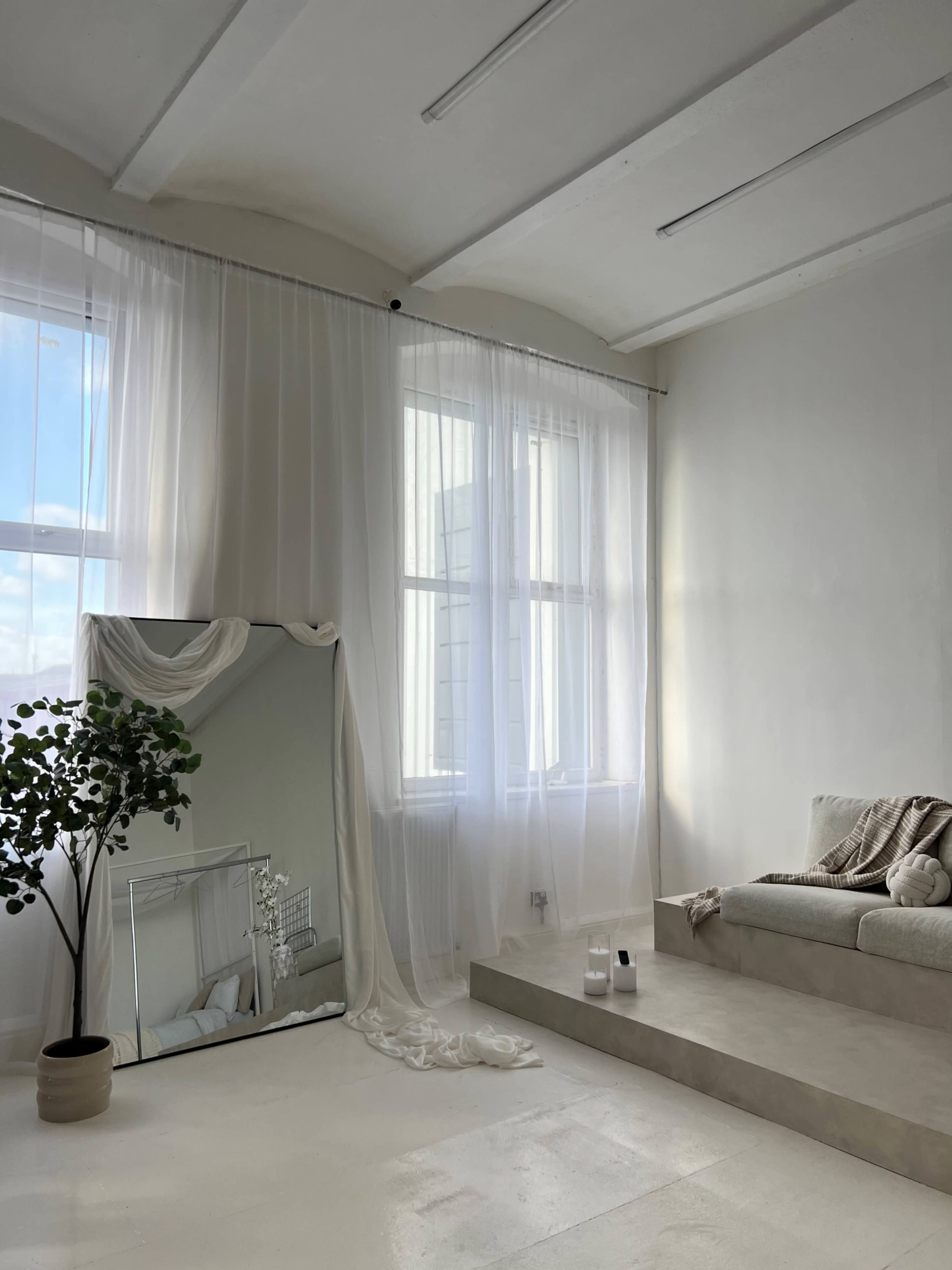 The image features a minimalist living room with a large mirror, sheer white curtains, a light-colored sofa, and a plant in a pot.