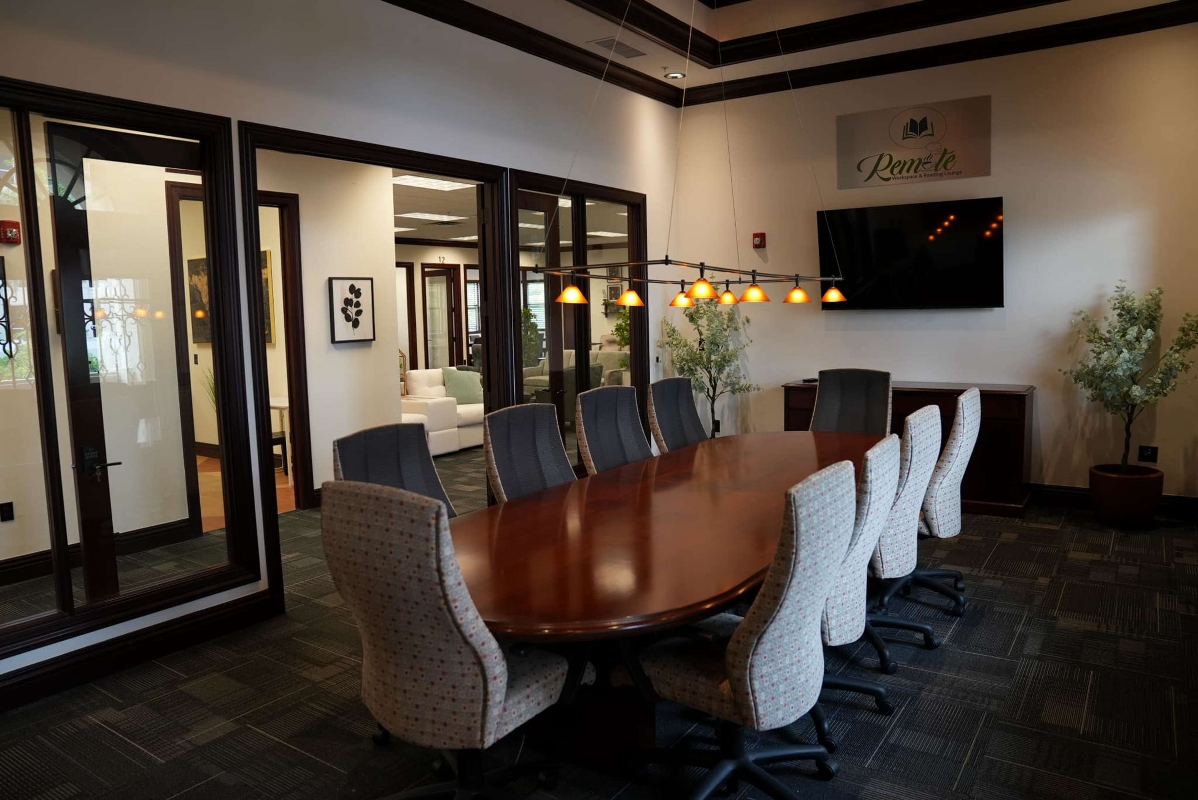 The image displays a modern conference room featuring a long wooden table surrounded by upholstered chairs, with decorative lighting overhead and a television mounted on the wall.