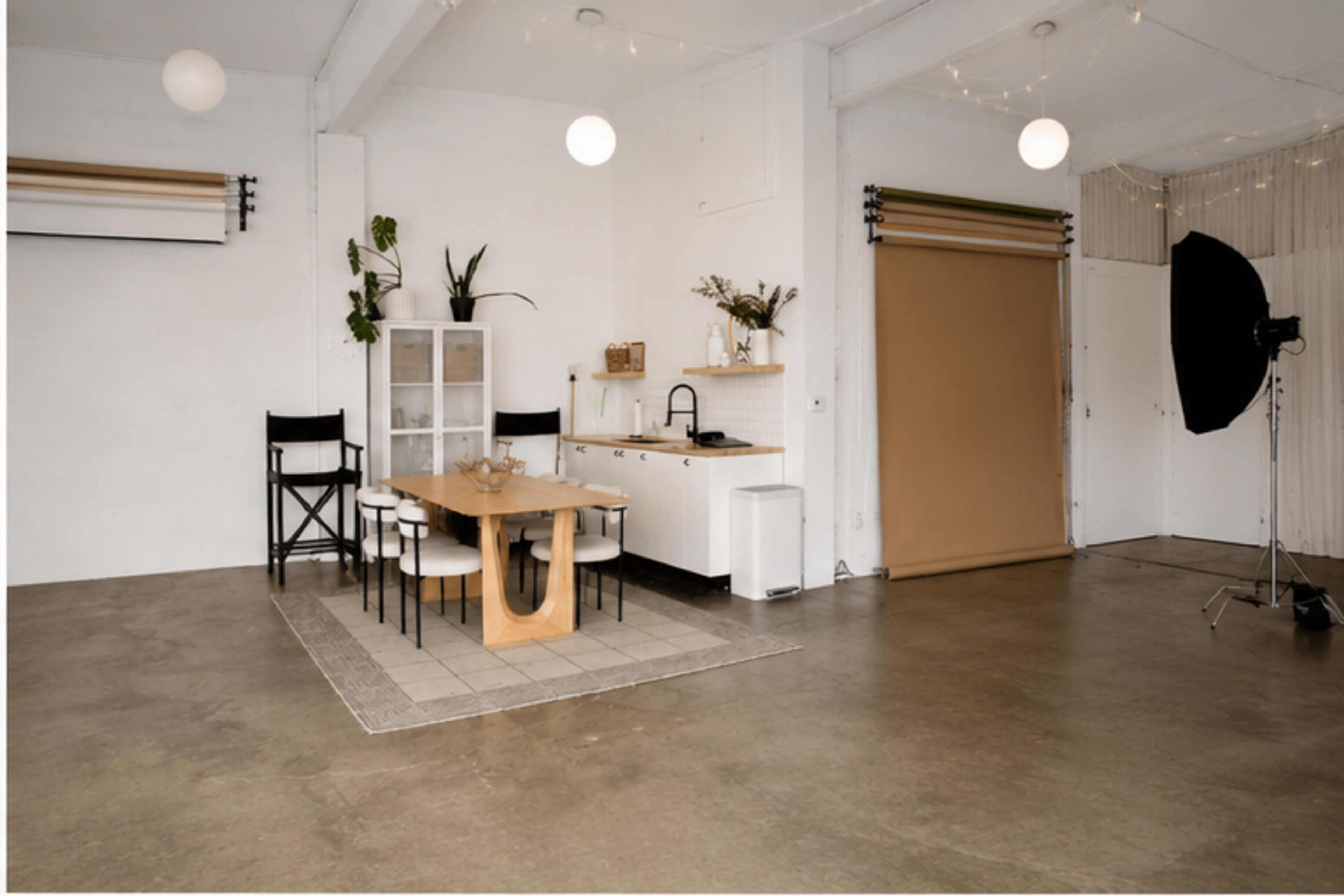 The image shows a minimalist kitchen area with a wooden dining table, black chairs, and a backdrop for photography.