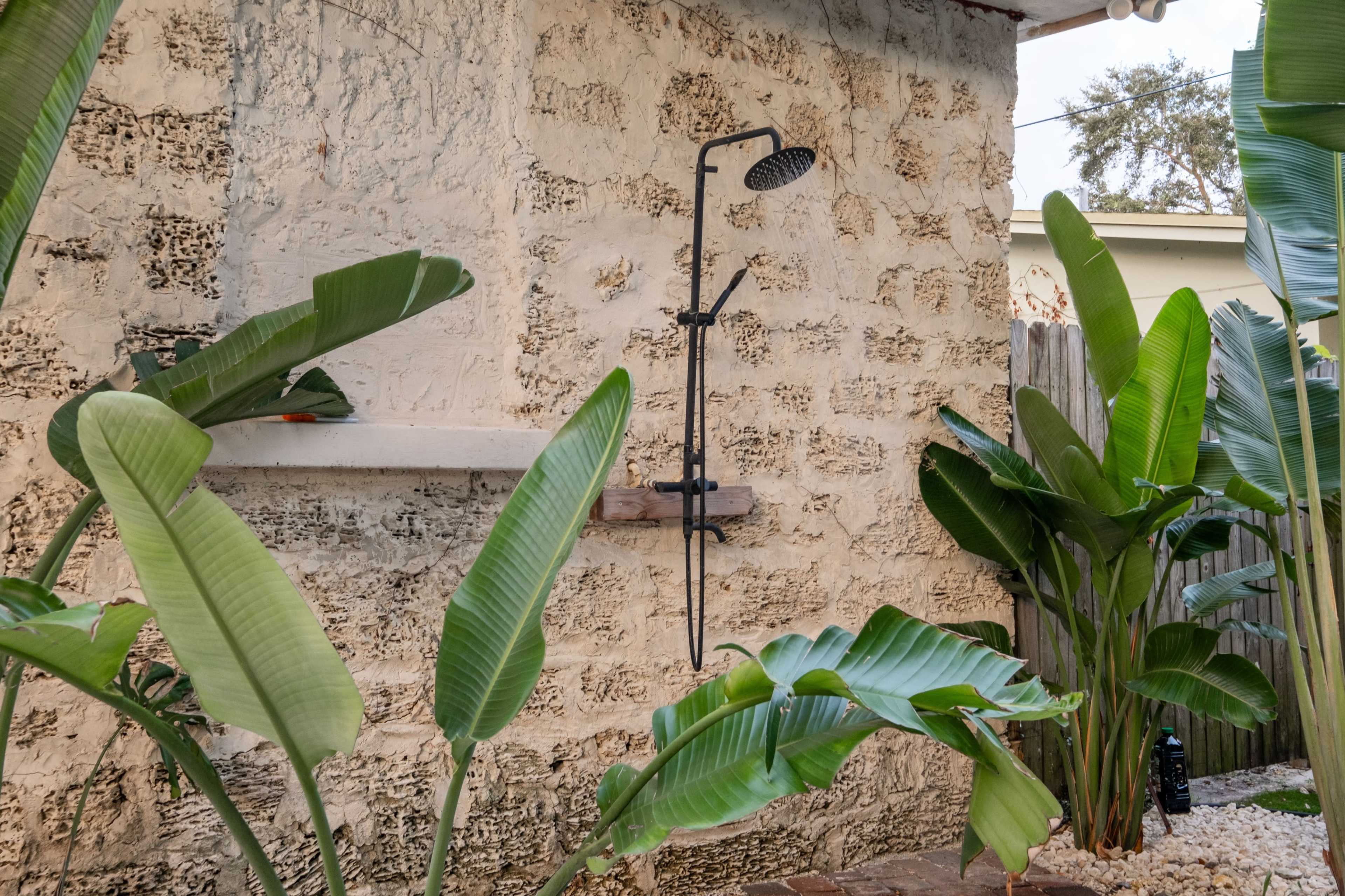 An outdoor shower is mounted on a textured wall, surrounded by large green plants.
