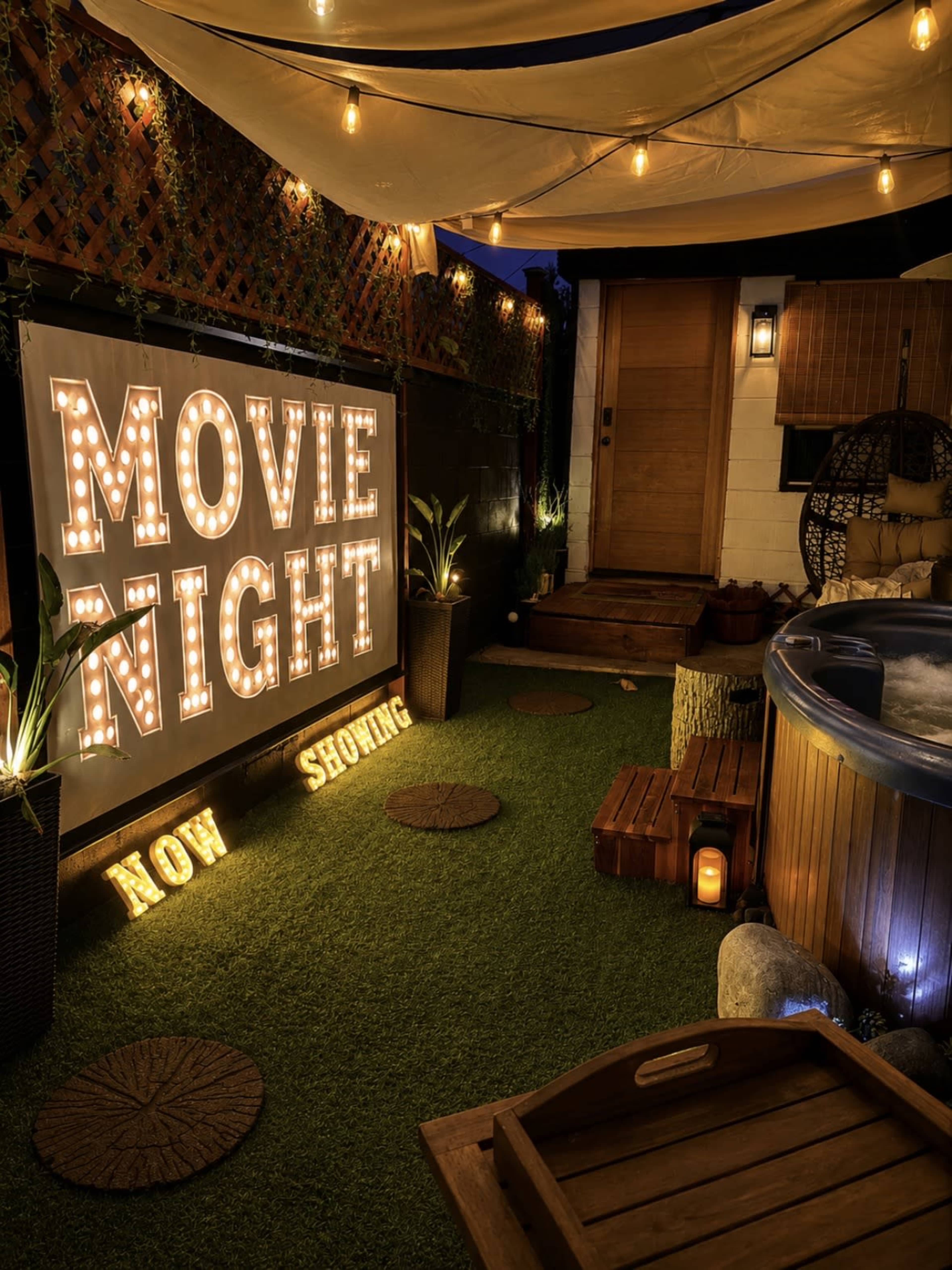 A cozy outdoor space is decorated with a large illuminated sign reading "MOVIE NIGHT NOW SHOWING," surrounded by plants, a hot tub, and soft lighting.