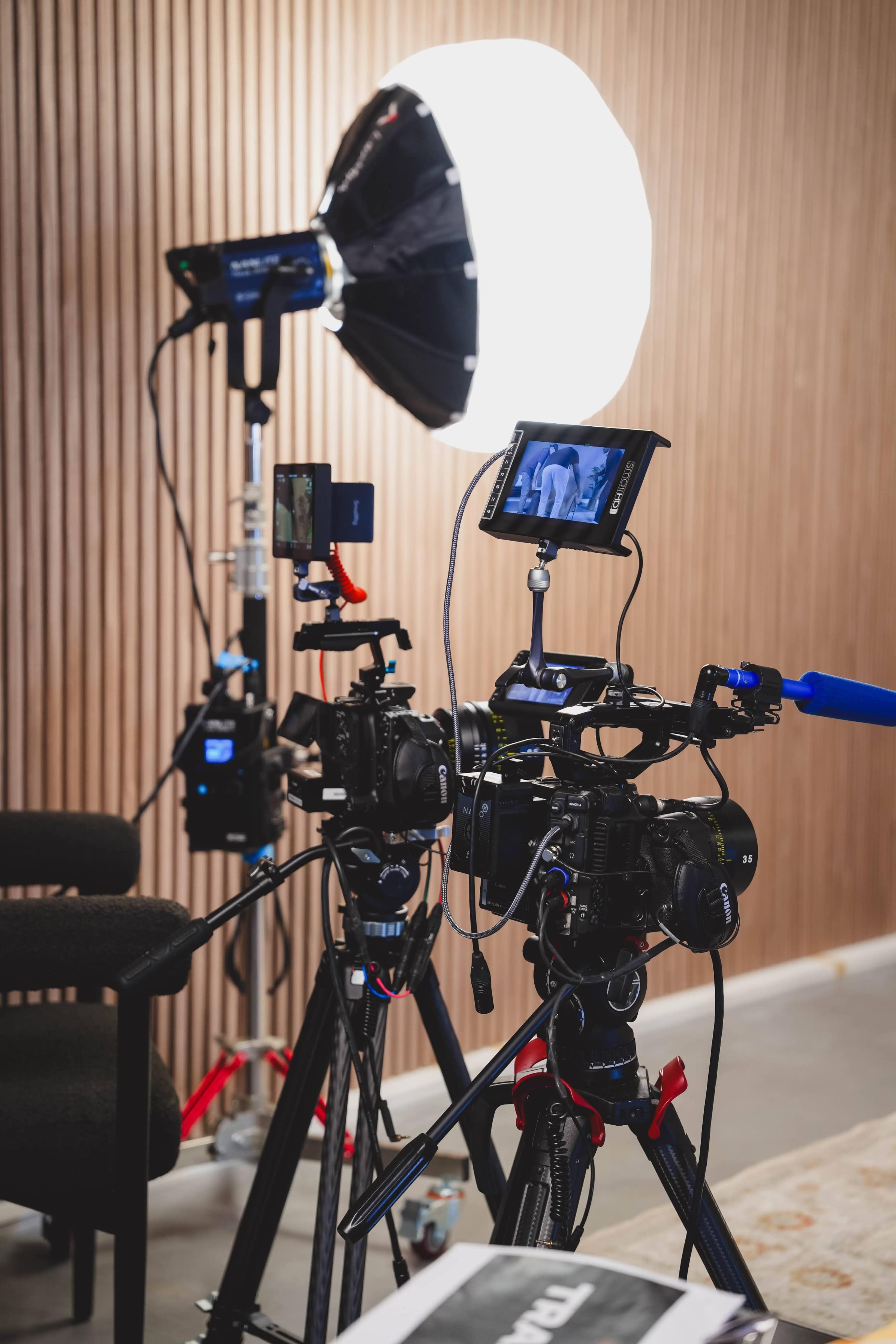 The image shows two video cameras set on tripods, positioned next to a large softbox light in a studio setting with wooden paneling in the background.