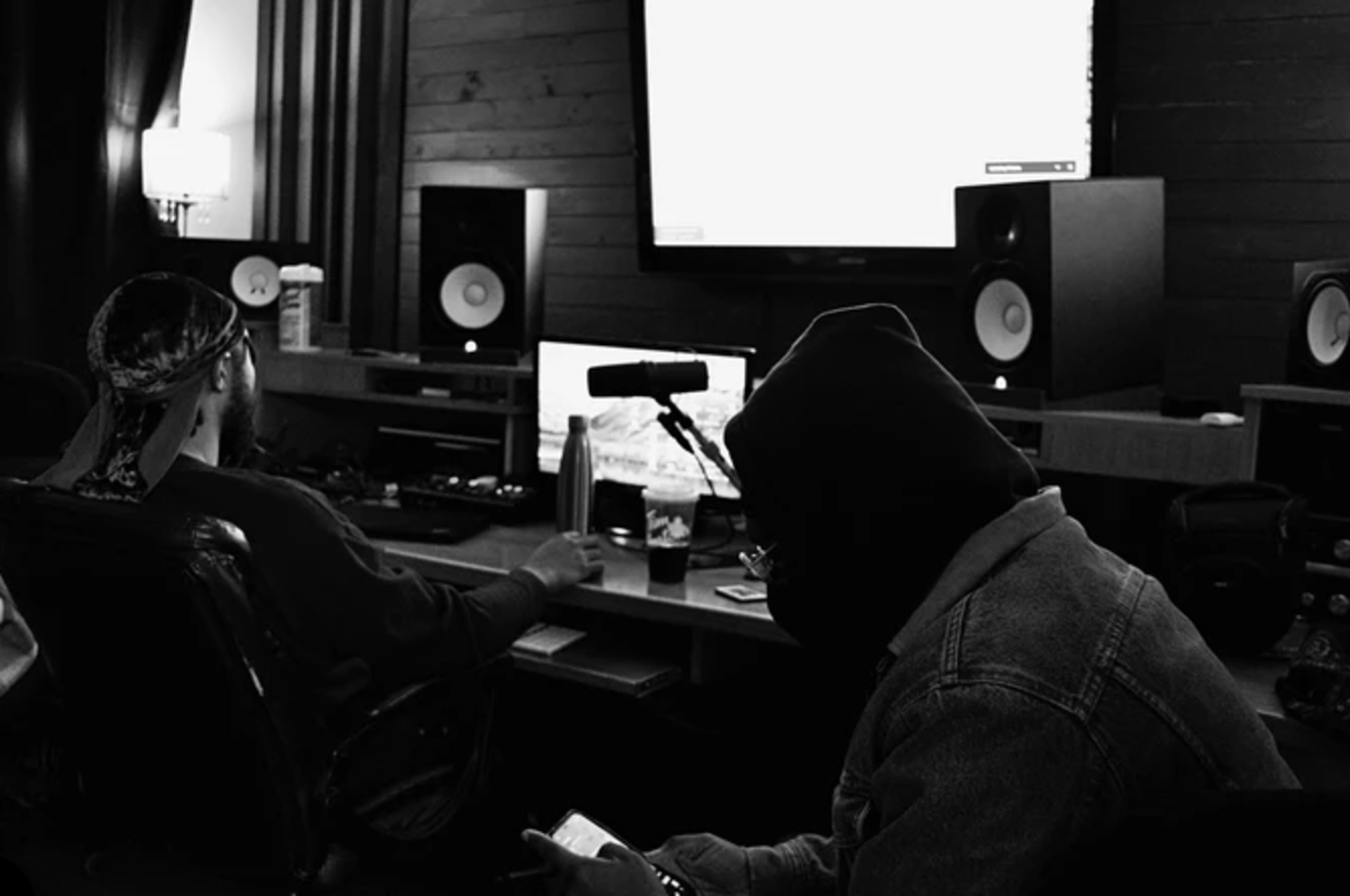Two individuals are working in a music studio, engaged with a computer and audio equipment while a microphone is visible on the desk.