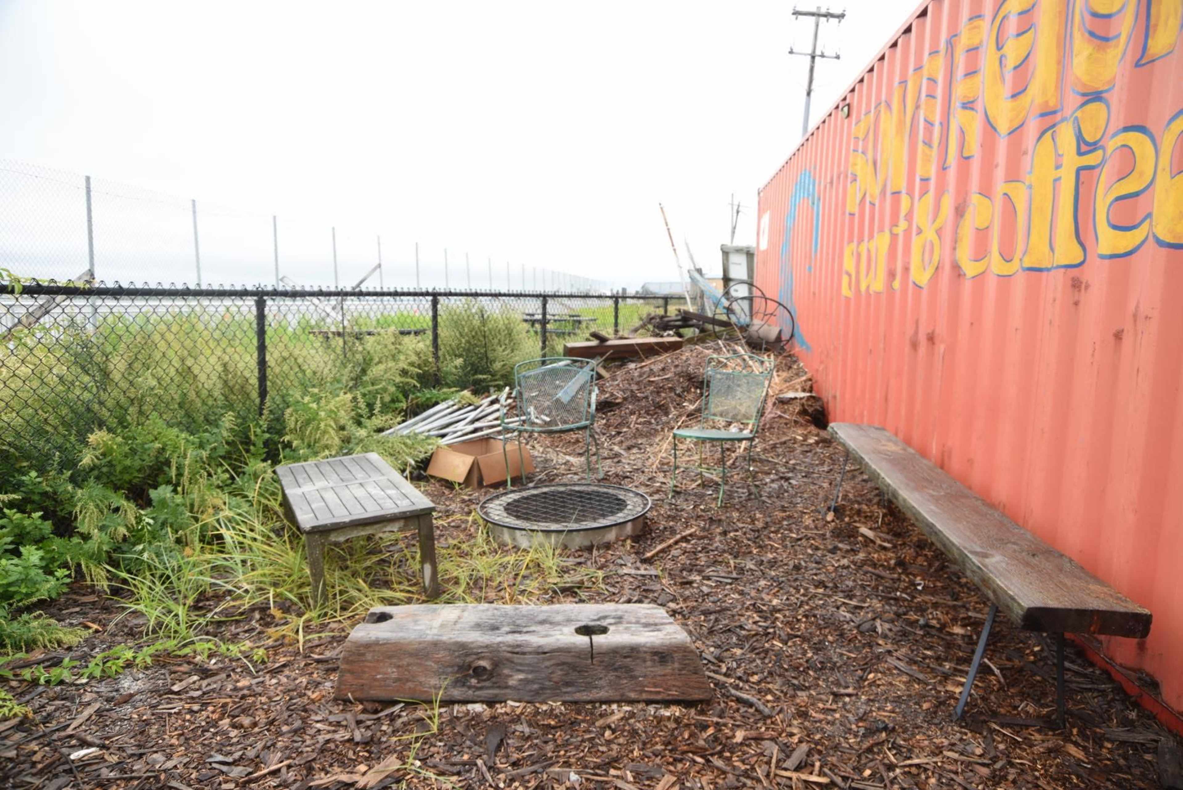 The scene shows an outdoor area beside a brightly painted shipping container, featuring wooden benches, metal chairs, and scattered debris among the overgrown vegetation.