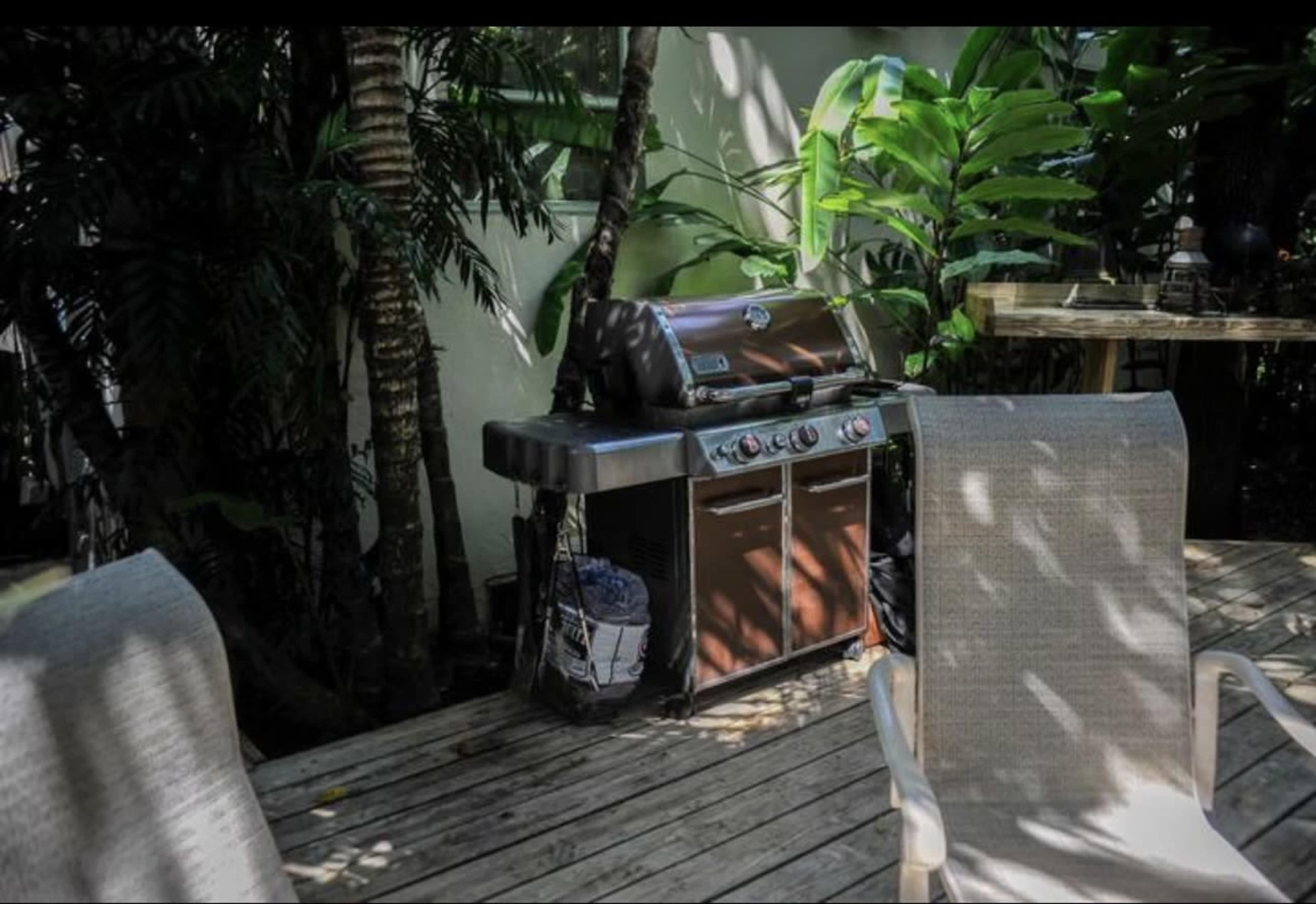 A barbecue grill is placed on a wooden deck surrounded by dense greenery and potted plants.