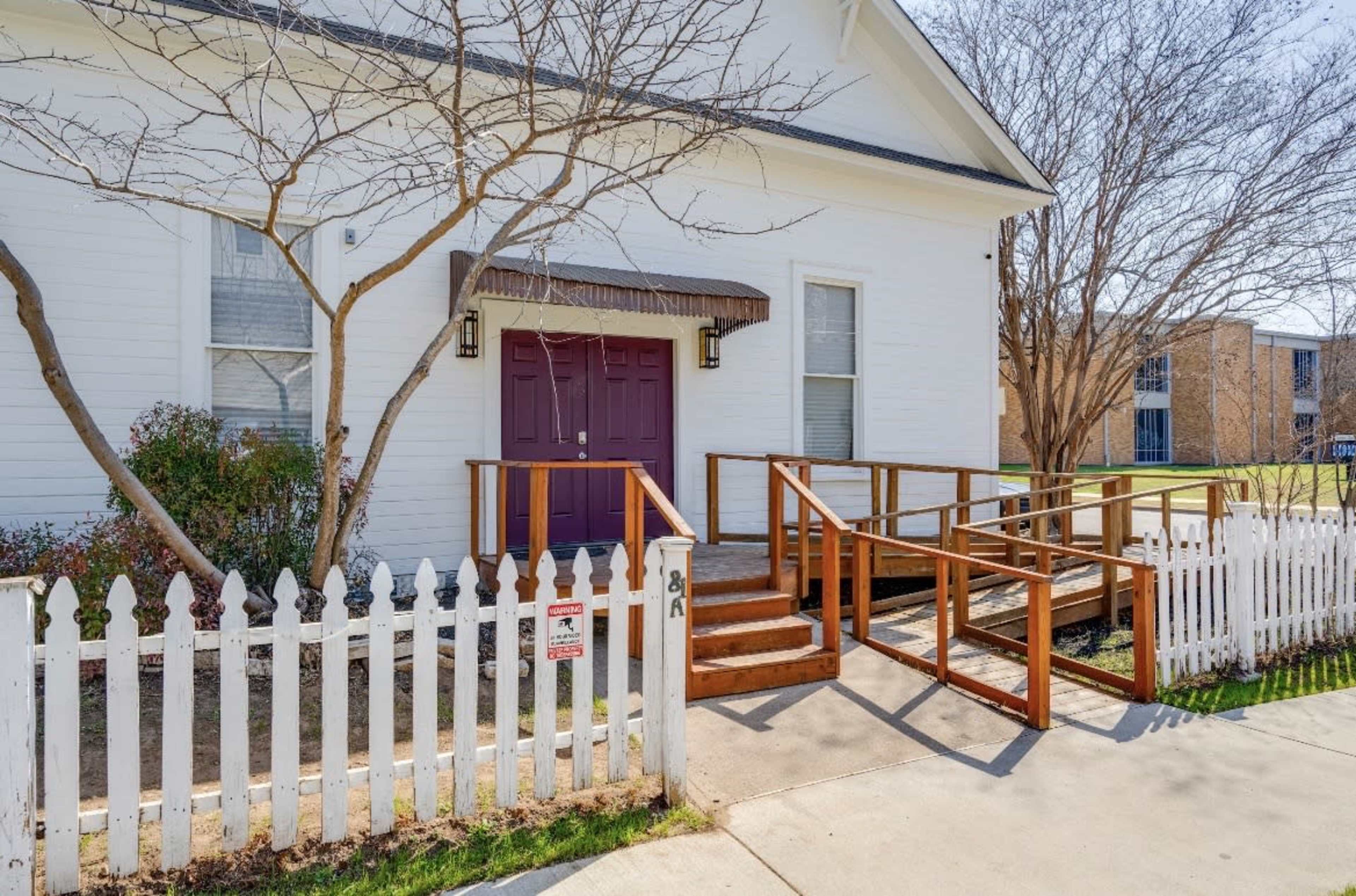 The image shows a white building with a purple double door, a wooden ramp, and a white picket fence surrounding the front yard.