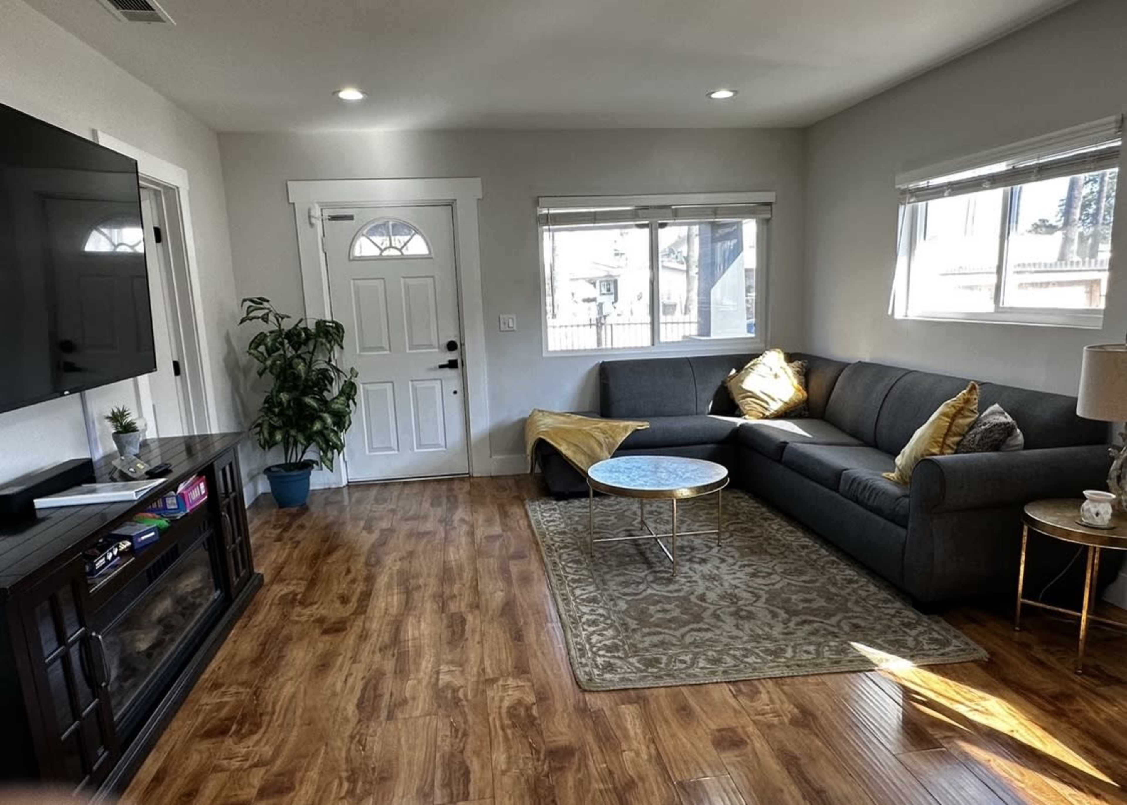 The room features a gray sectional sofa, a small coffee table, a television mounted on the wall, and a front door leading outside.