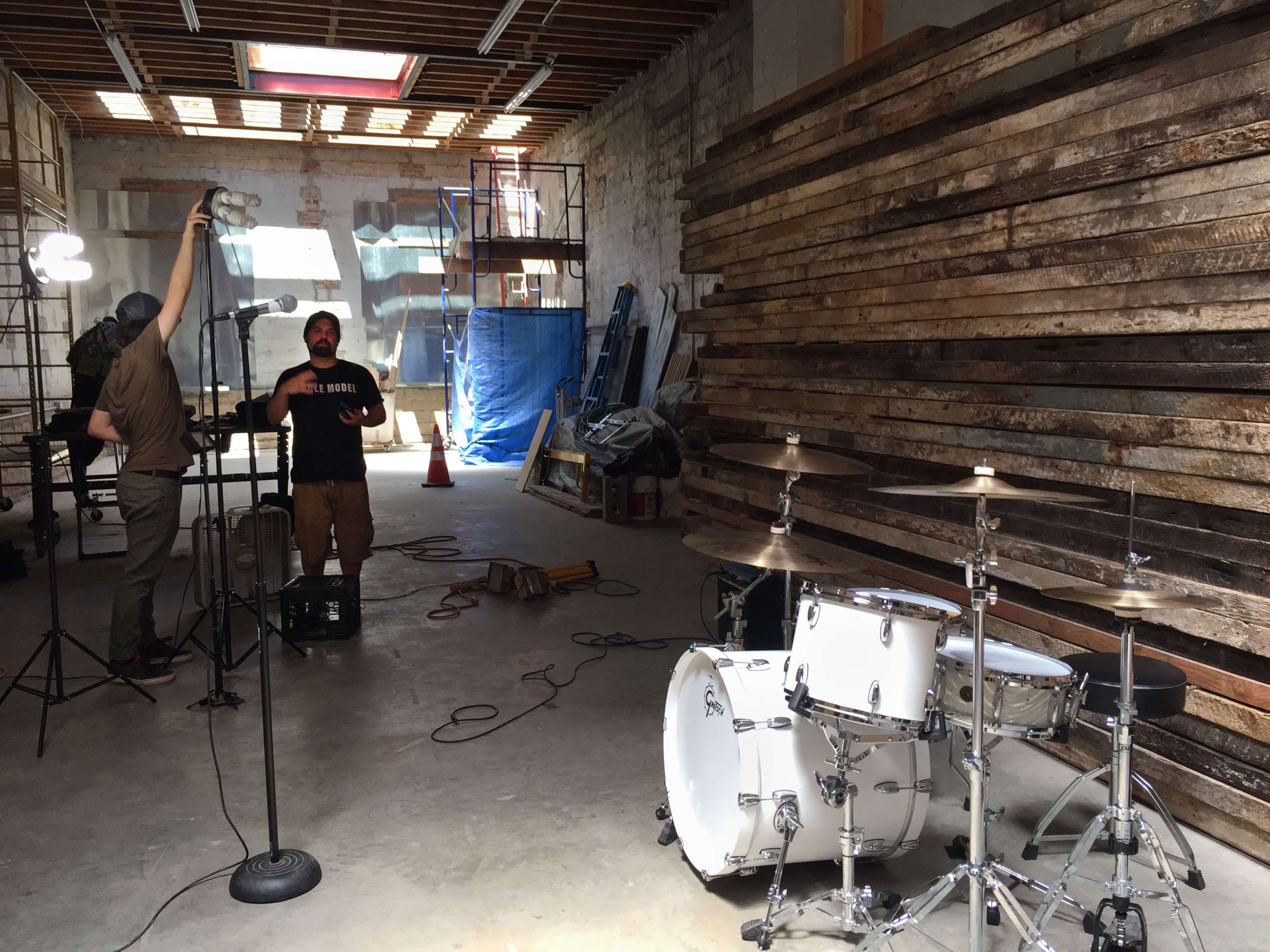 A musician stands with a microphone in a spacious, unfinished warehouse, next to a drum set, while a crew member adjusts lighting equipment.