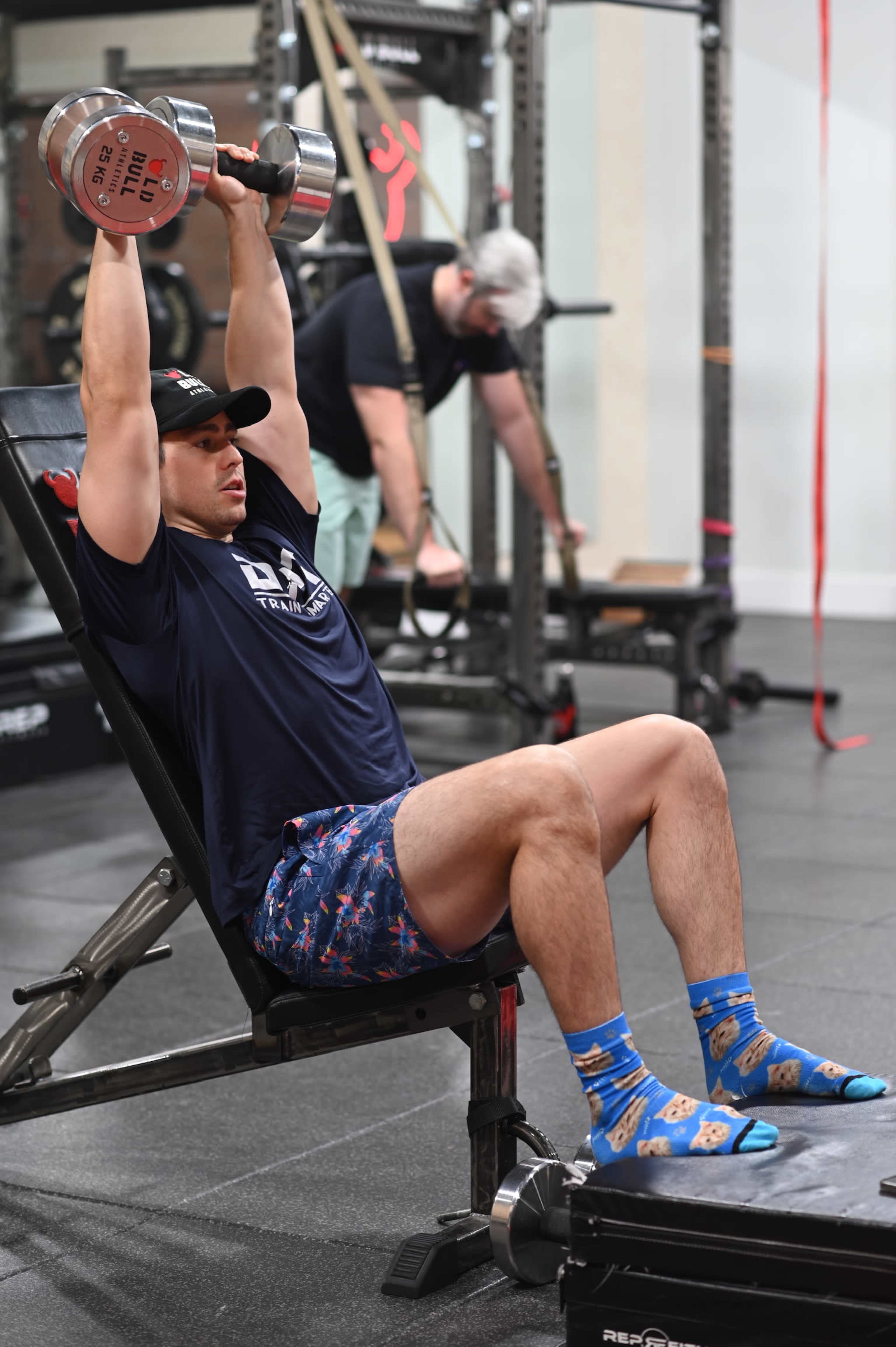 A man performs a seated overhead dumbbell press in a gym while another person adjusts equipment in the background.