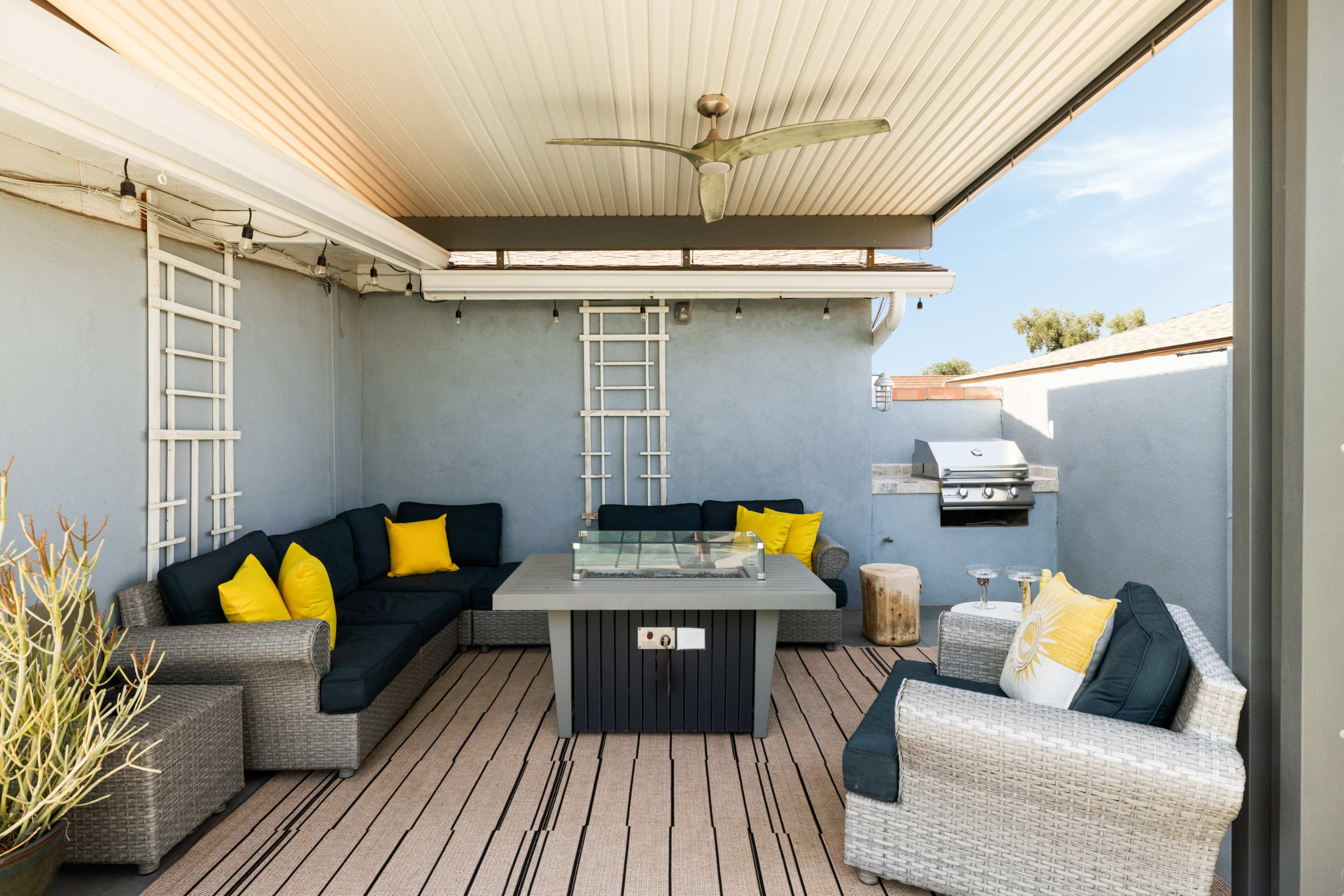 A shaded outdoor seating area features a sectional sofa with yellow accent pillows, a central coffee table, and a grill on the side.