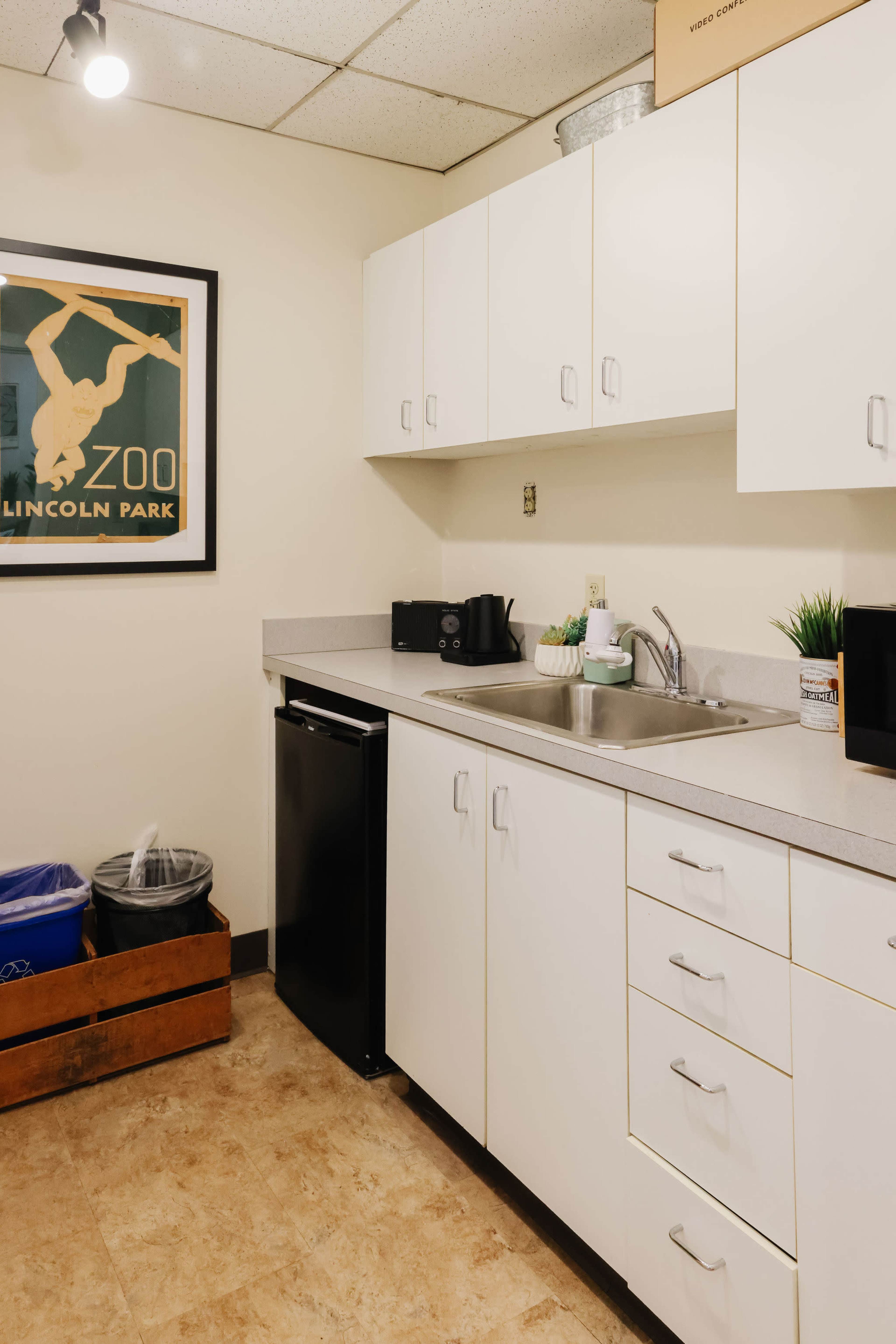 The image shows a small kitchen area featuring a sink, black refrigerator, and white cabinets, alongside a wall poster and waste bins.