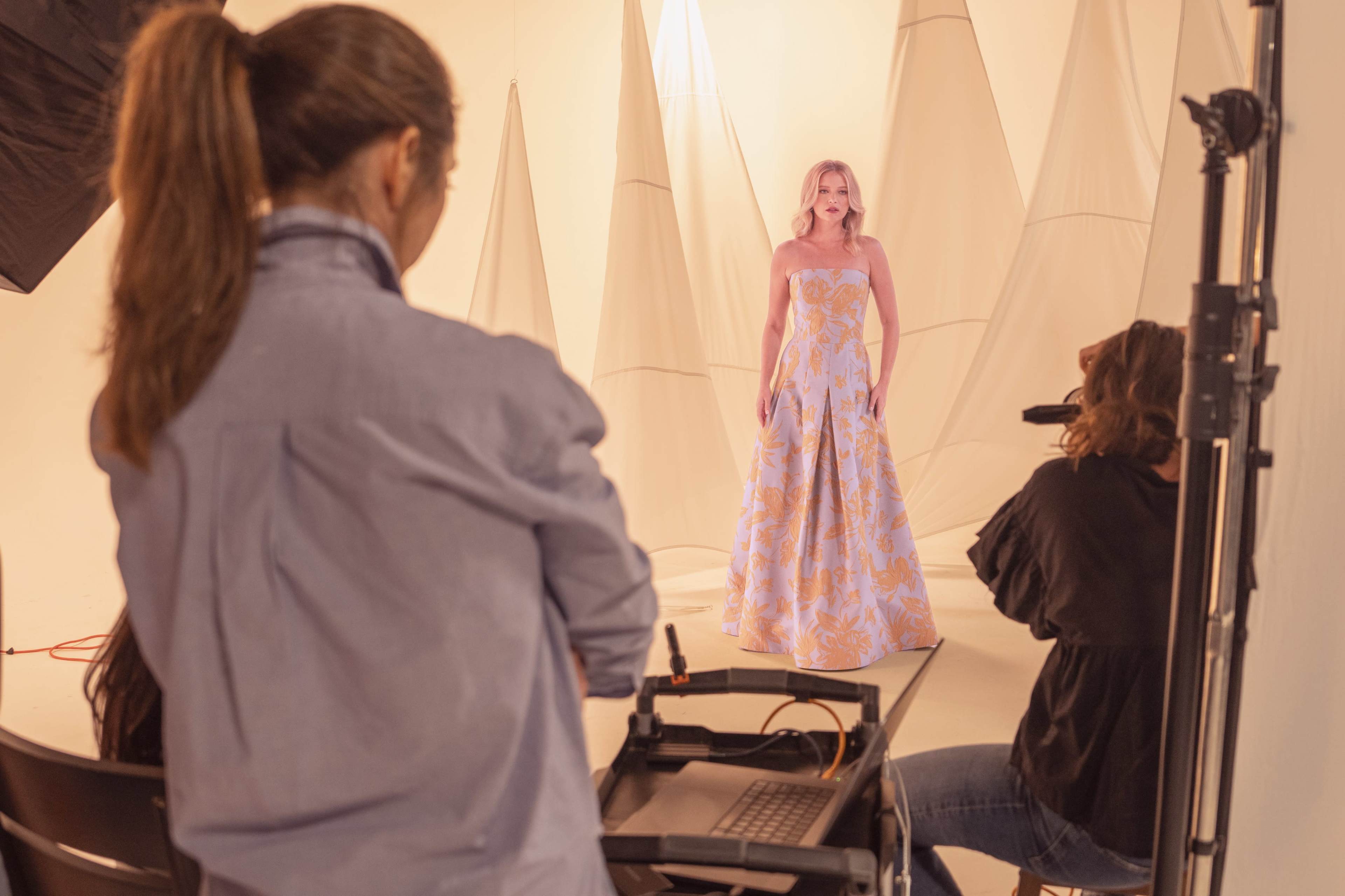 A model in a floral dress poses for a photoshoot in a studio with fabric backdrops.