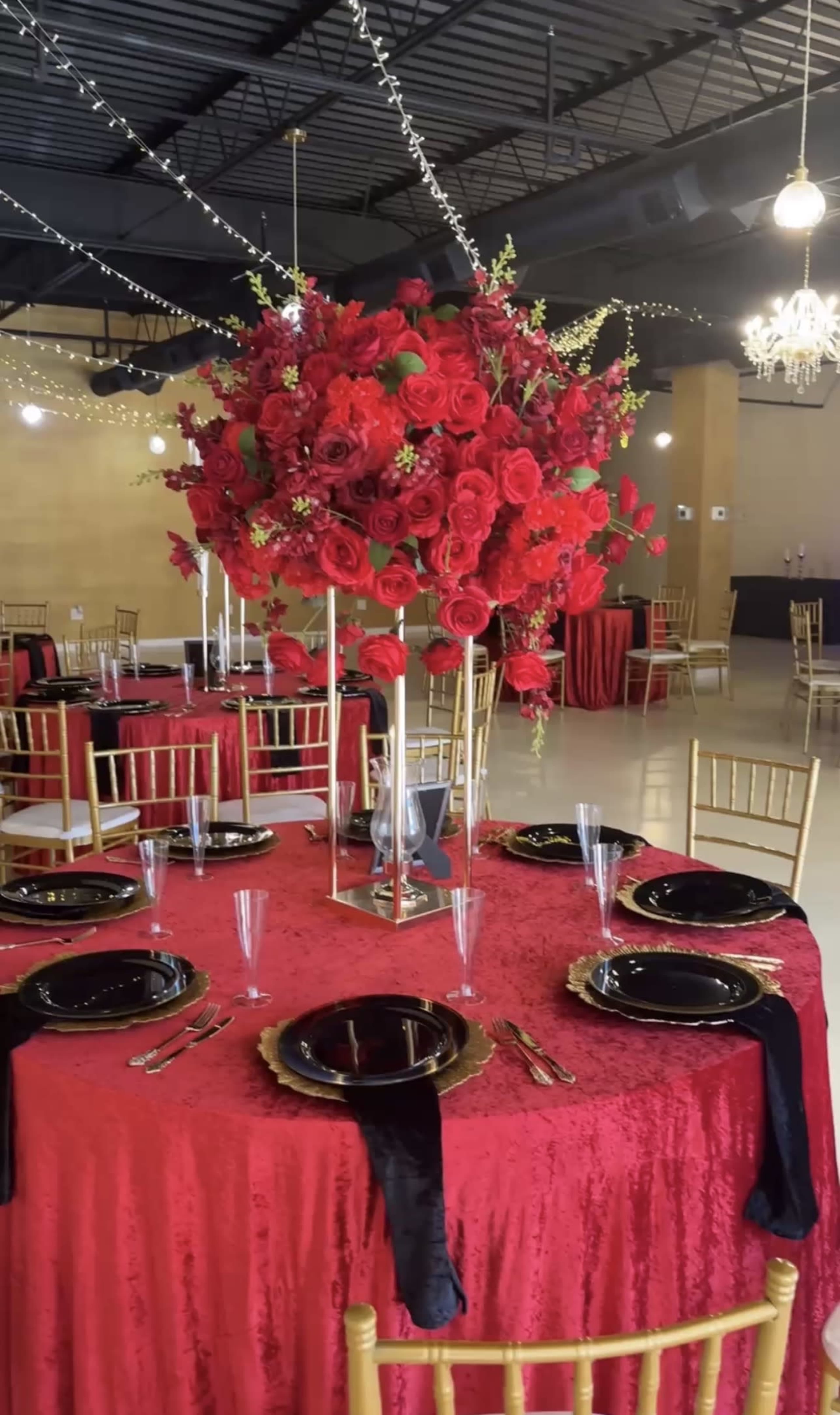 A lavish dining area is set with gold-framed chairs and elegantly arranged tables draped in red fabric, featuring tall centerpieces of vibrant red flowers and black plates.